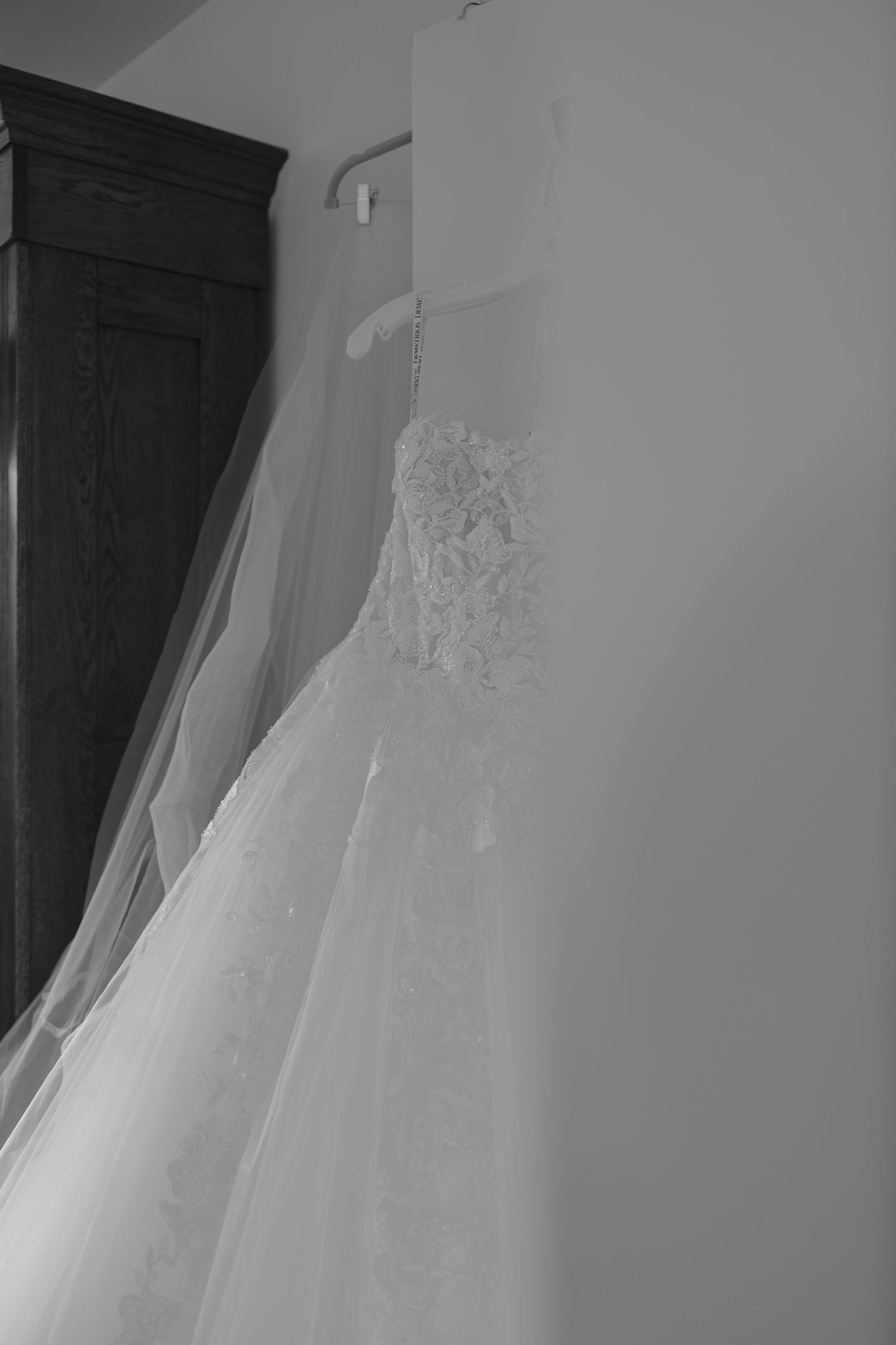 Black and white photo of an elegant lace wedding dress hanging on a wall hanger near a wooden armoire.