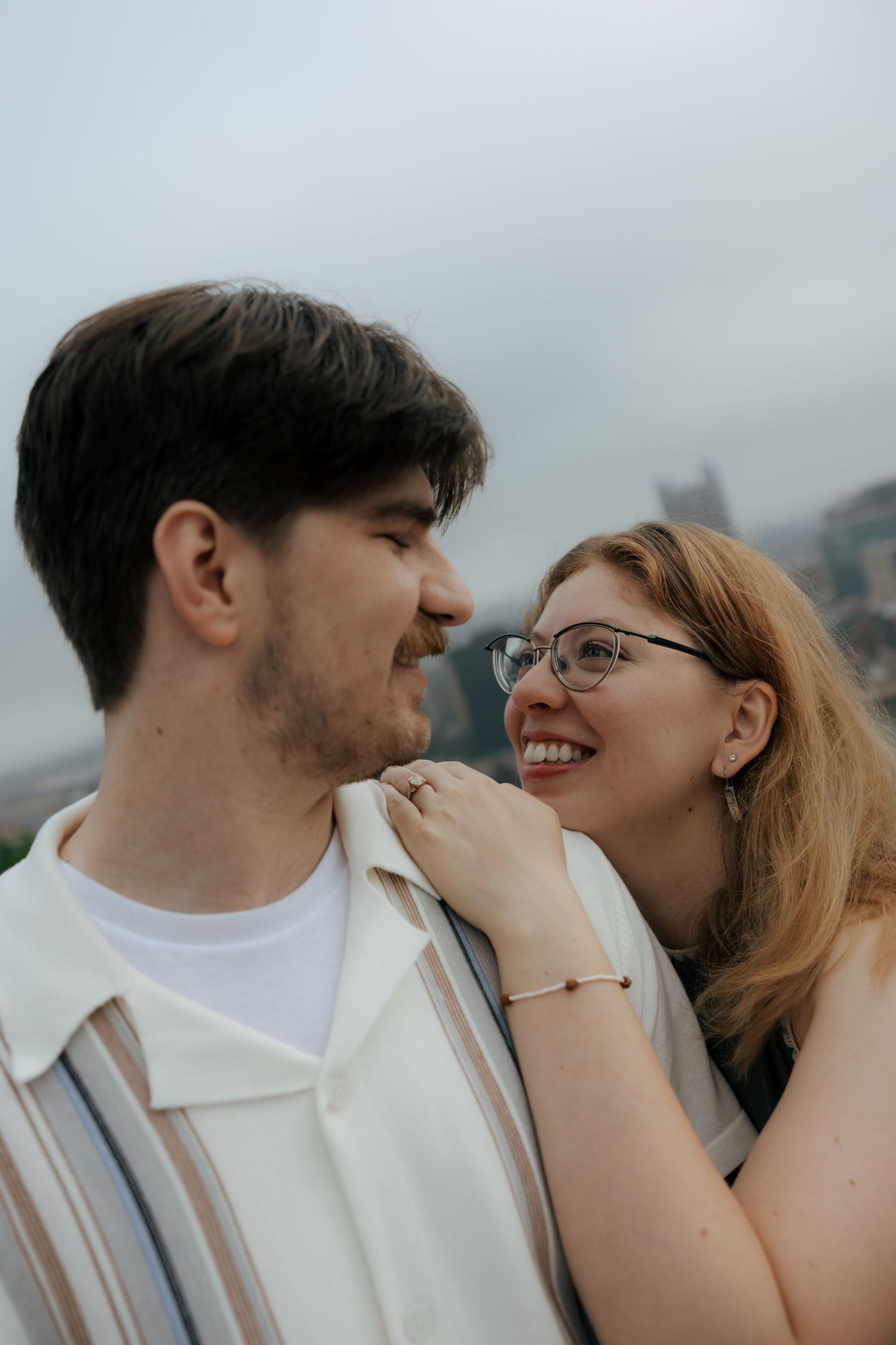 A smiling woman with glasses leans close to her partner outdoors with a city skyline in the background.