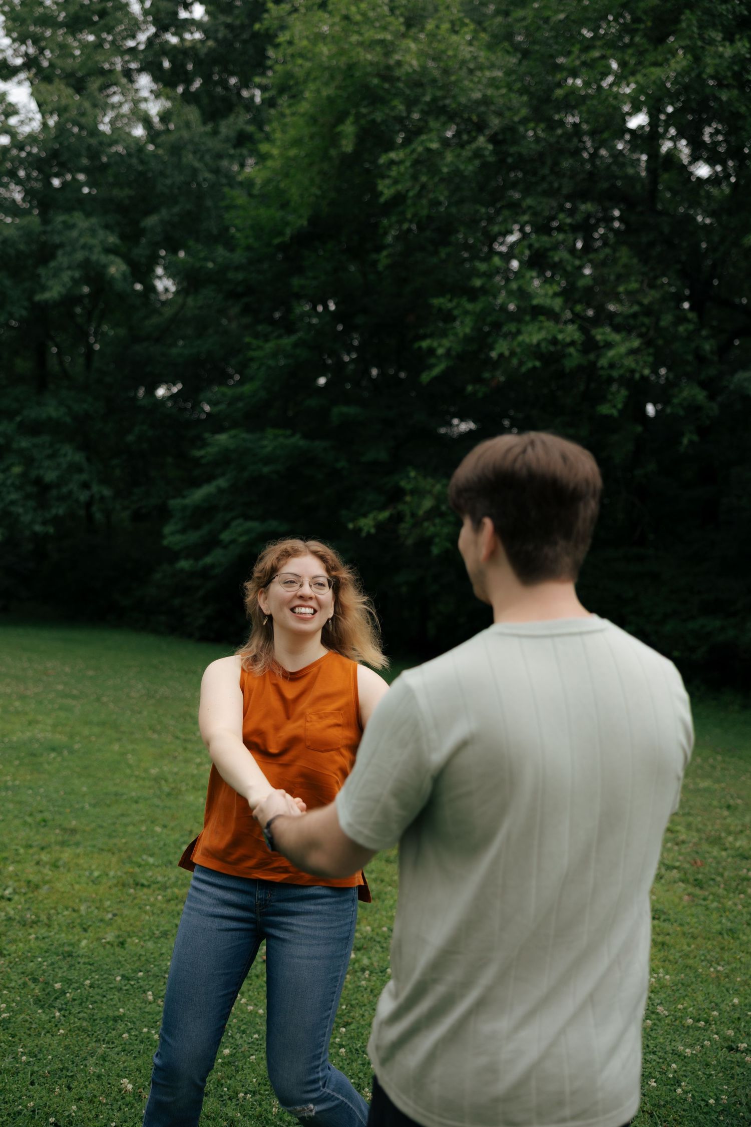 A smiling woman in an orange sleeveless top holds hands with a man on a green grassy field.