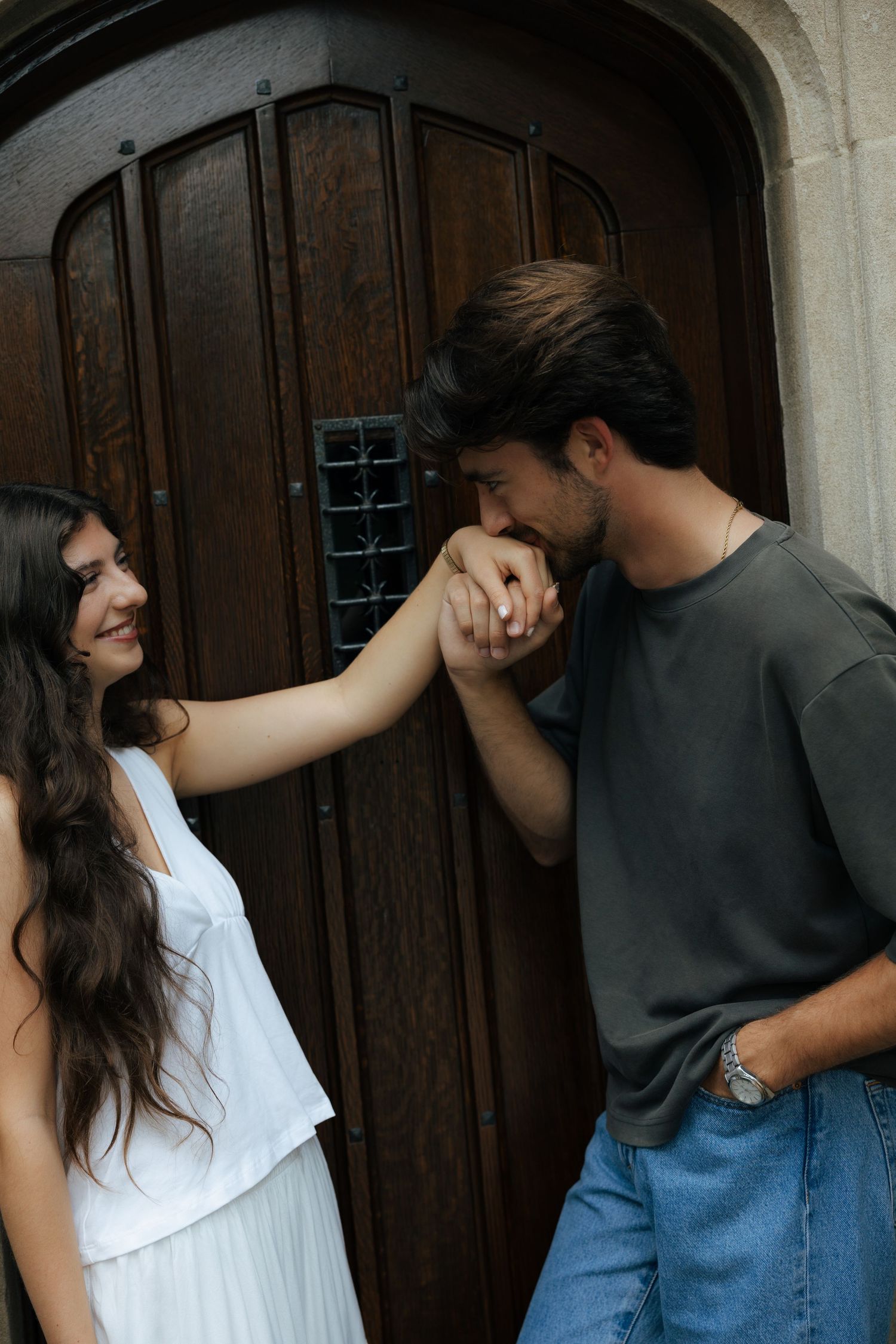 A man gallantly kisses a woman's hand in front of a grand wooden arched door.