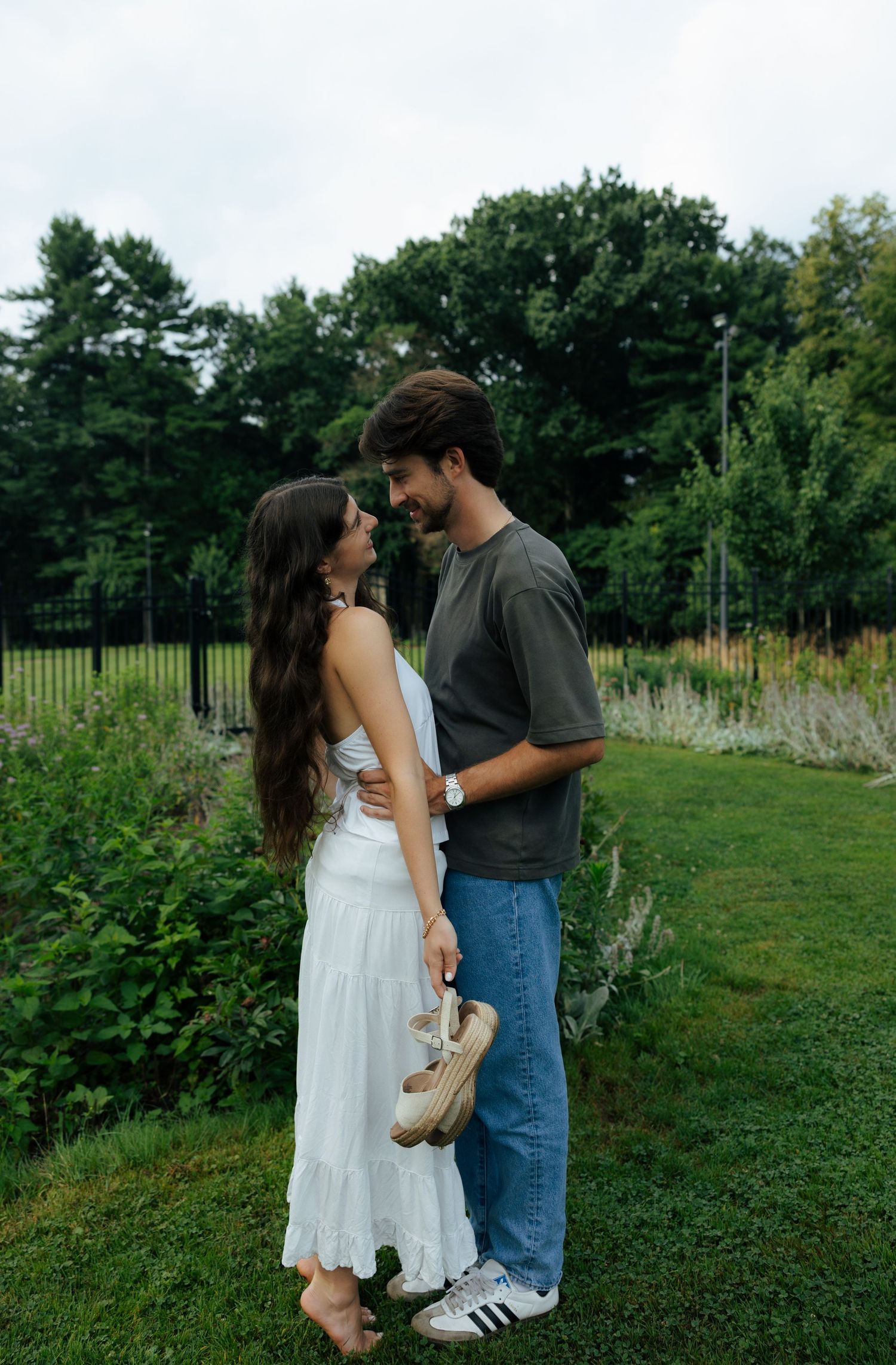 A couple stands face to face smiling on a green lawn, the man holding sandals while surrounded by lush greenery.