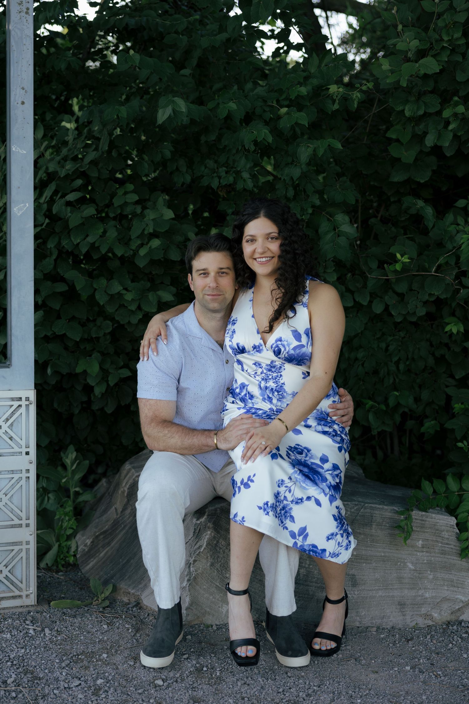 A smiling couple poses together on a stone bench with green trees in the background.