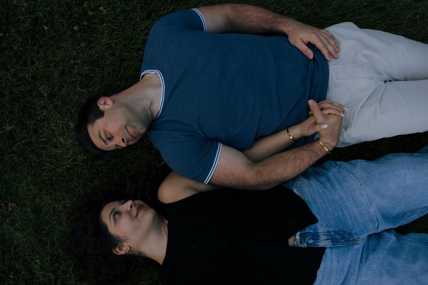 Couple lying on grass holding hands, viewed from above, wearing casual blue and black outfits.