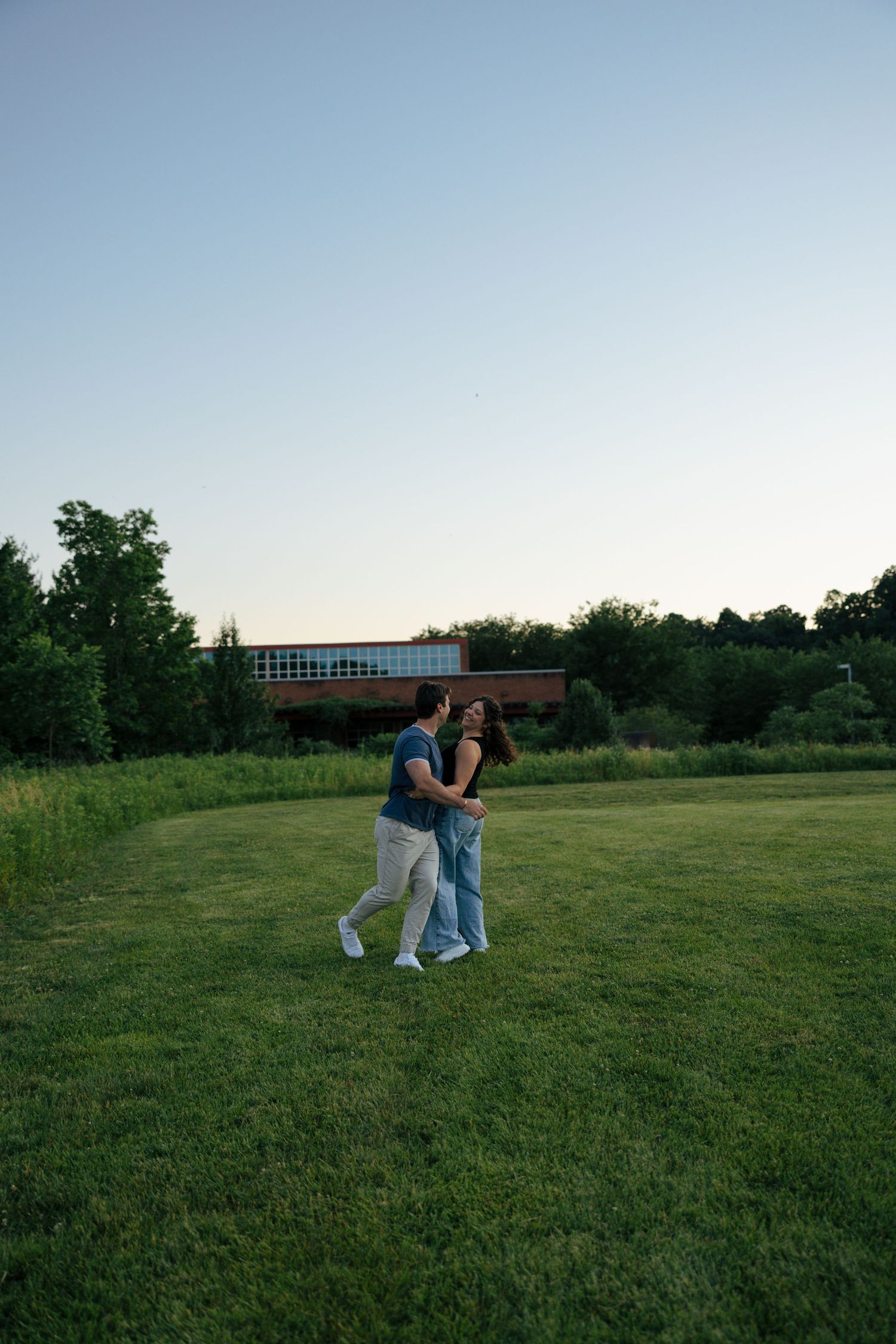 A couple embraces and dances together on a green lawn at dusk with trees and a building in the background.