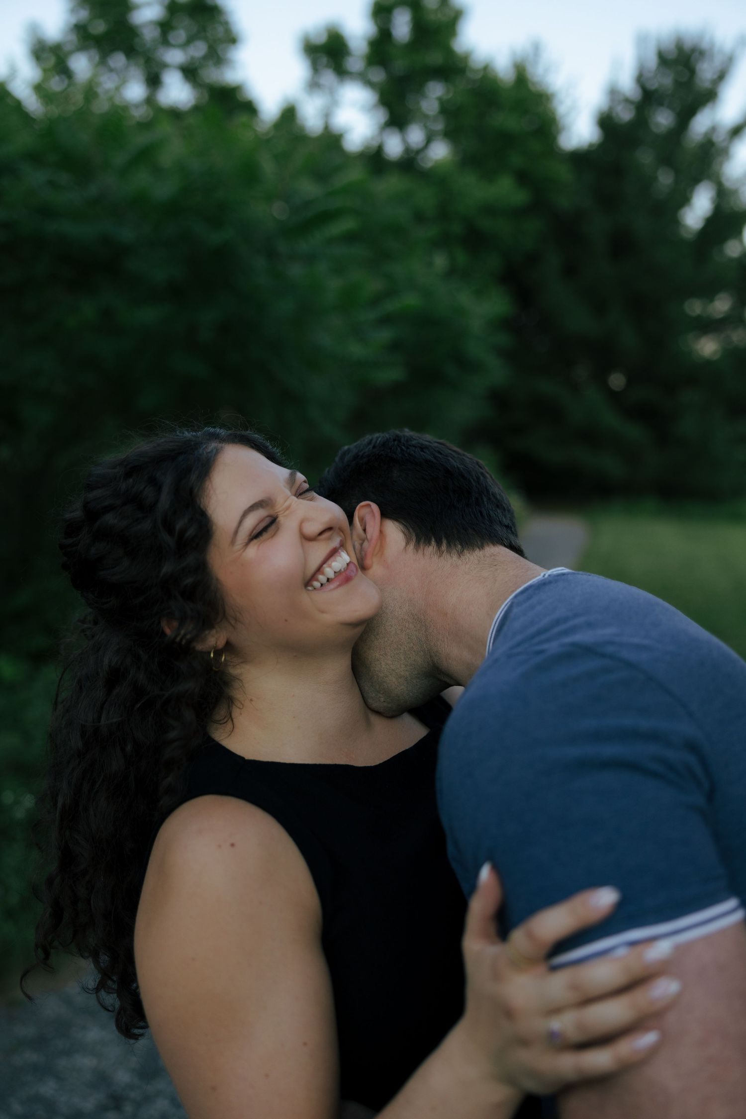 A woman laughs joyfully as her partner kisses her cheek outdoors with lush green trees in the background.