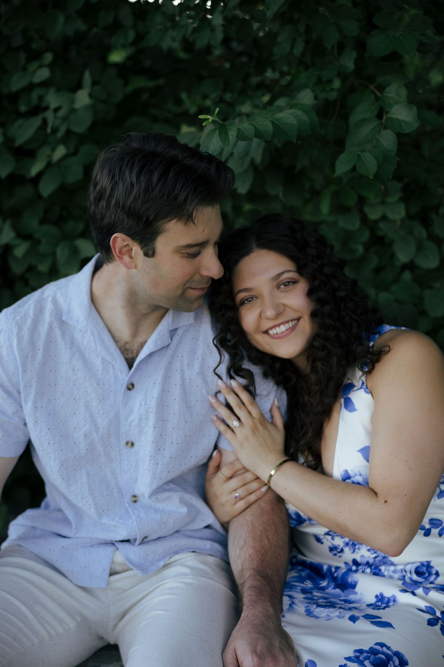 Smiling couple sitting together in front of lush green foliage, woman showing engagement ring.