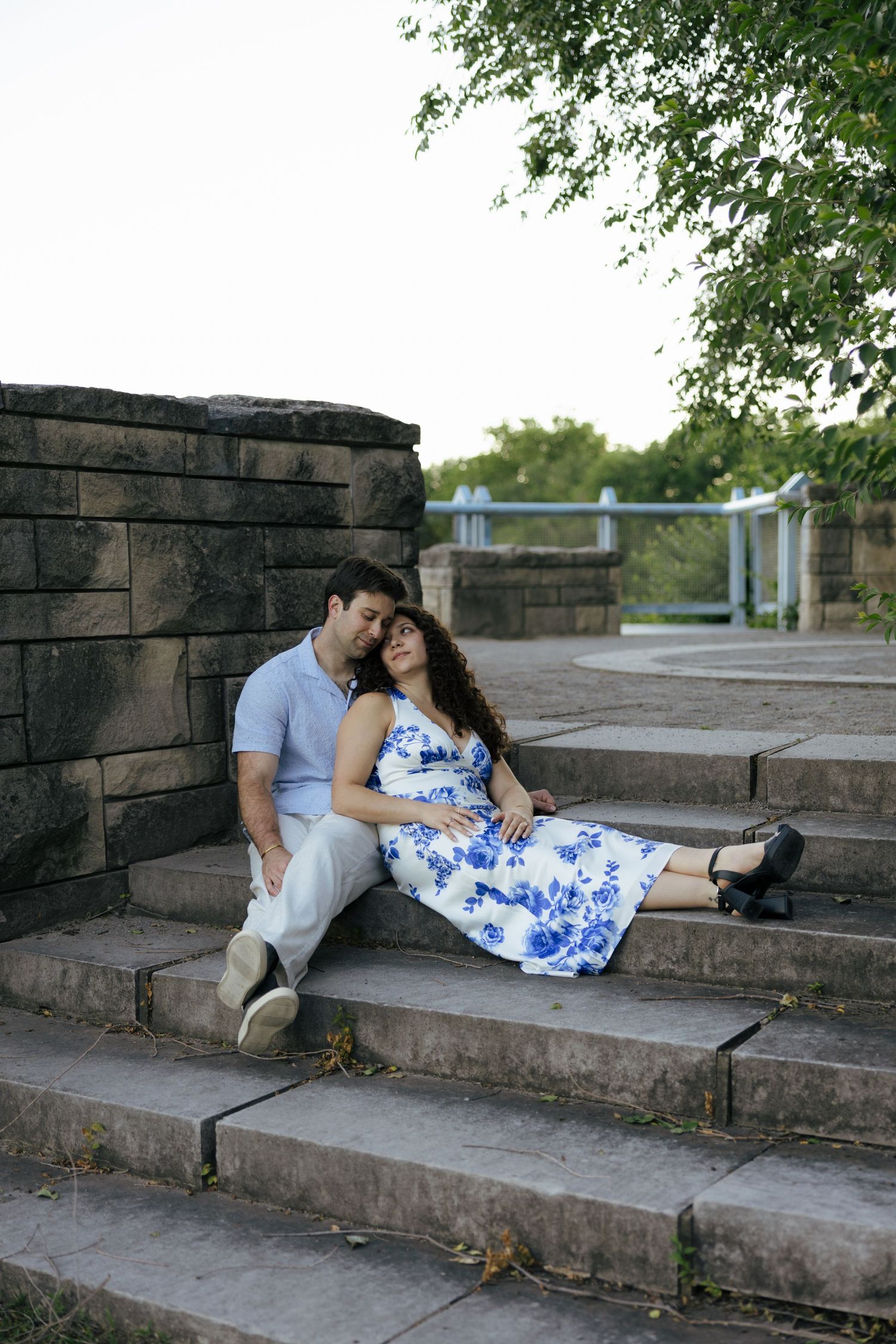 A couple relaxes on stone steps near a rustic wall, the woman in a blue floral dress leaning on her partner.
