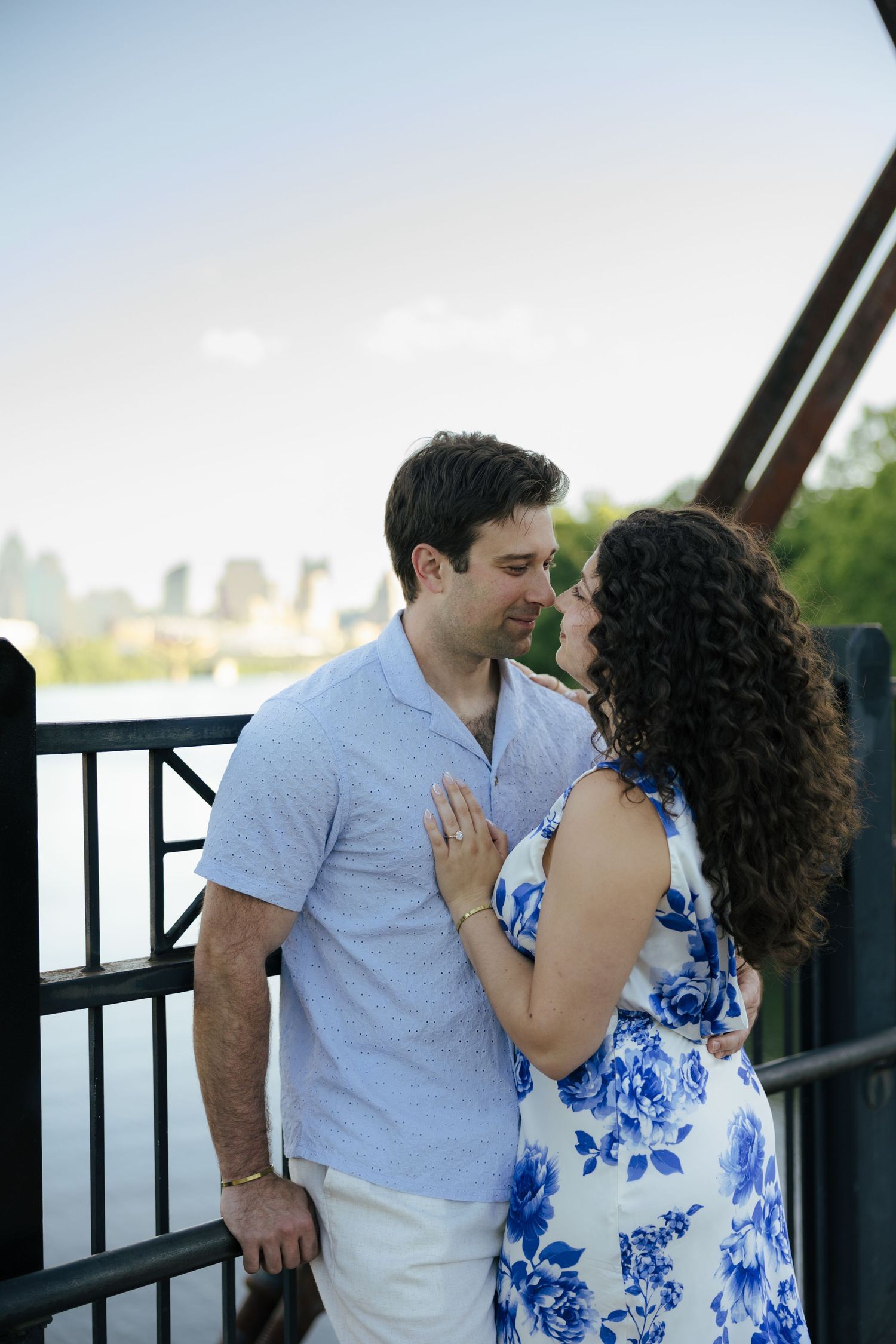 Couple nearly kissing on bridge with city skyline behind them, woman in blue floral dress.