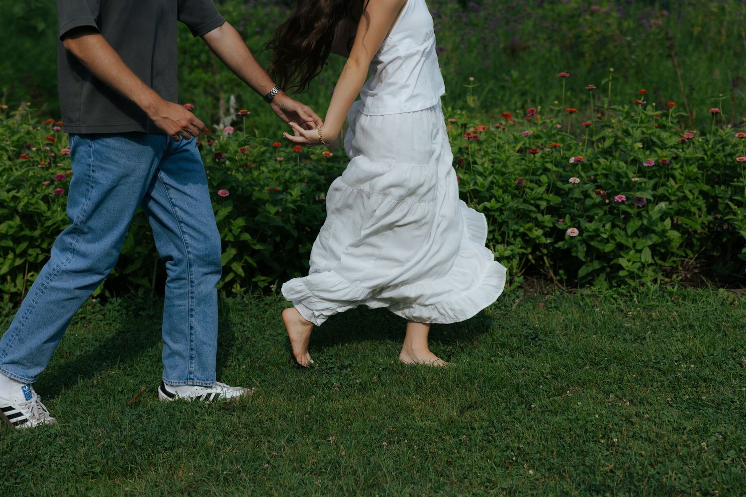 Couple holding hands running barefoot on green grass, woman in white dress mid-stride.