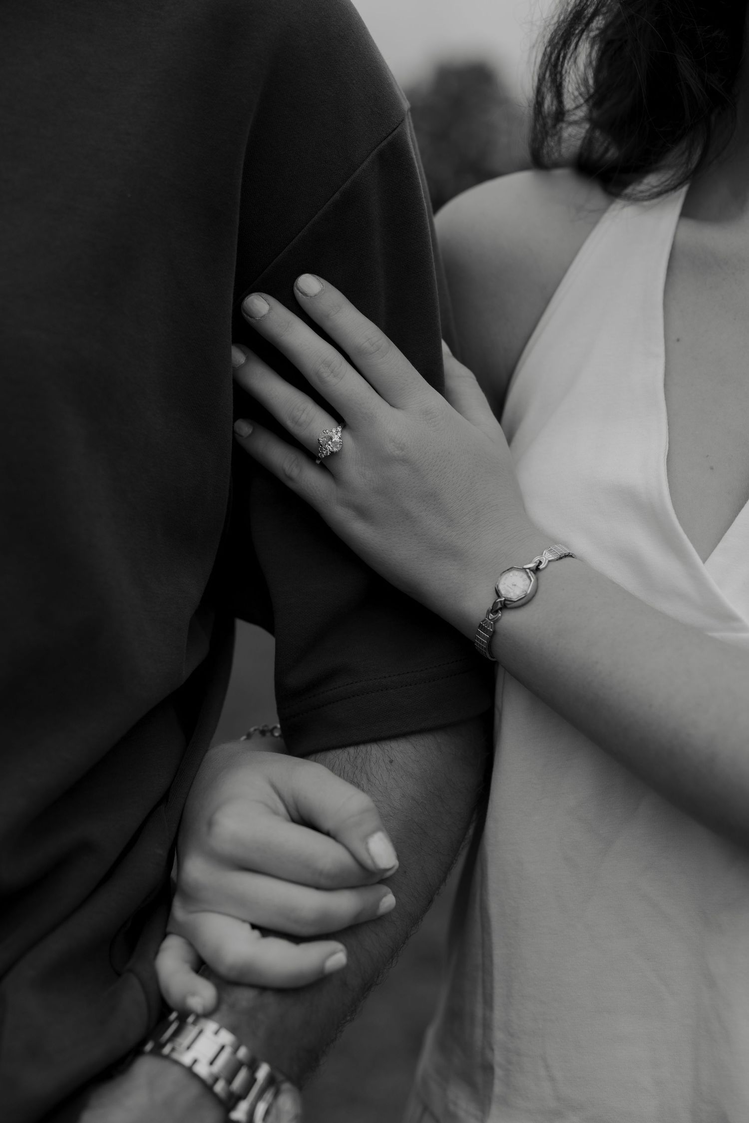 Black and white close-up of an engaged couples hands, woman displaying diamond ring while holding mans arm.
