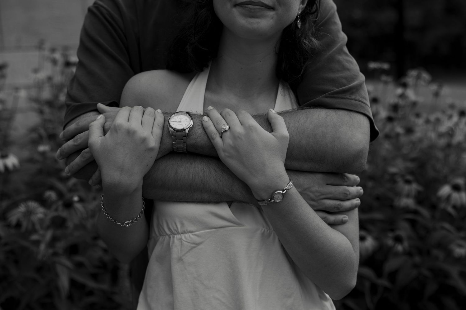 Black and white photo of a couple embracing, the man wrapping his arms around the woman from behind in a garden.