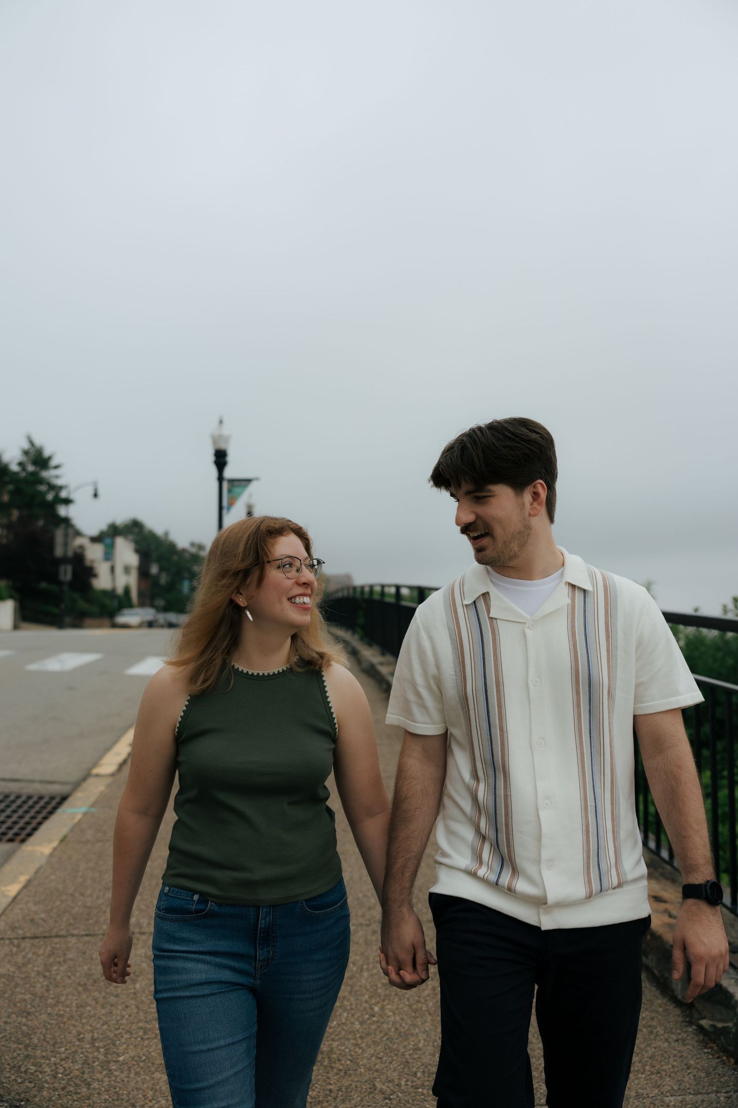 Couple holding hands and smiling at each other while walking along an urban waterfront promenade on a cloudy day.