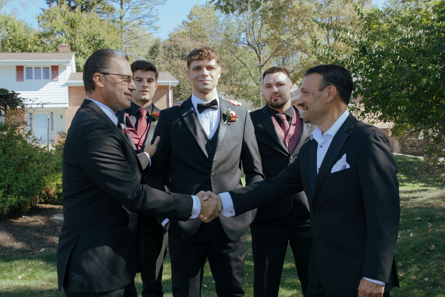 Men in formal suits shaking hands outdoors at a wedding, with a groom in a tan suit and boutonniere standing nearby.