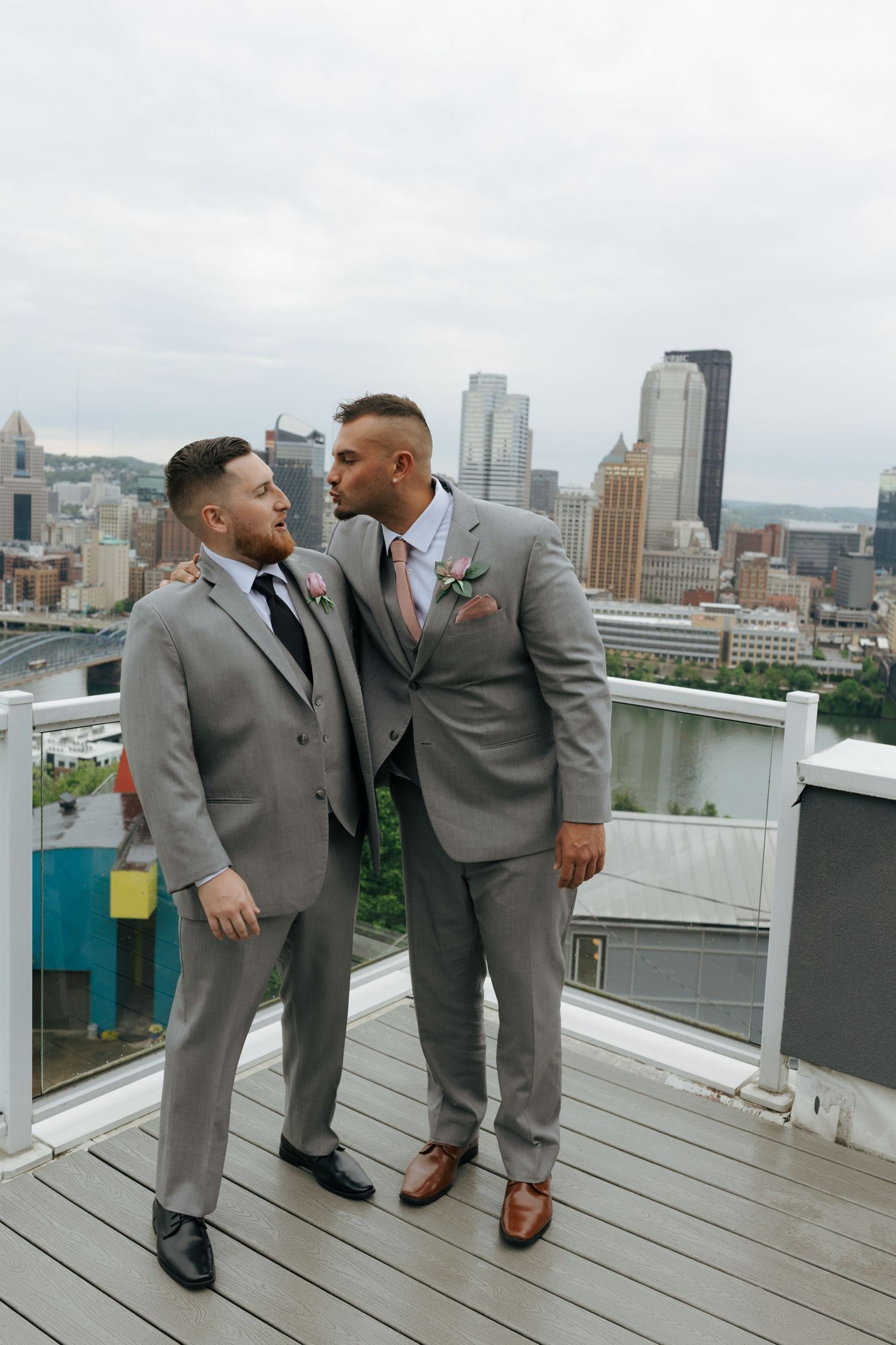 Two grooms in matching gray suits share a kiss on a rooftop deck with Pittsburgh city skyline backdrop.