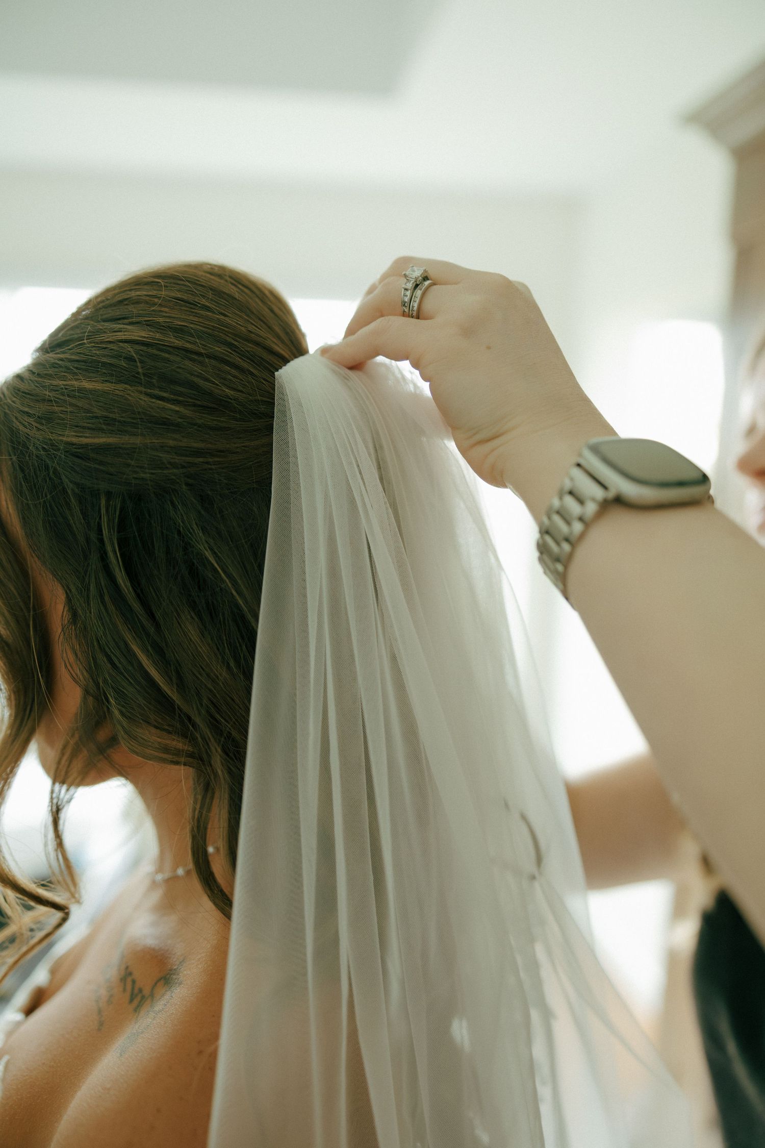 Assistant placing a delicate white bridal veil into bride's elegant updo hairstyle before wedding.