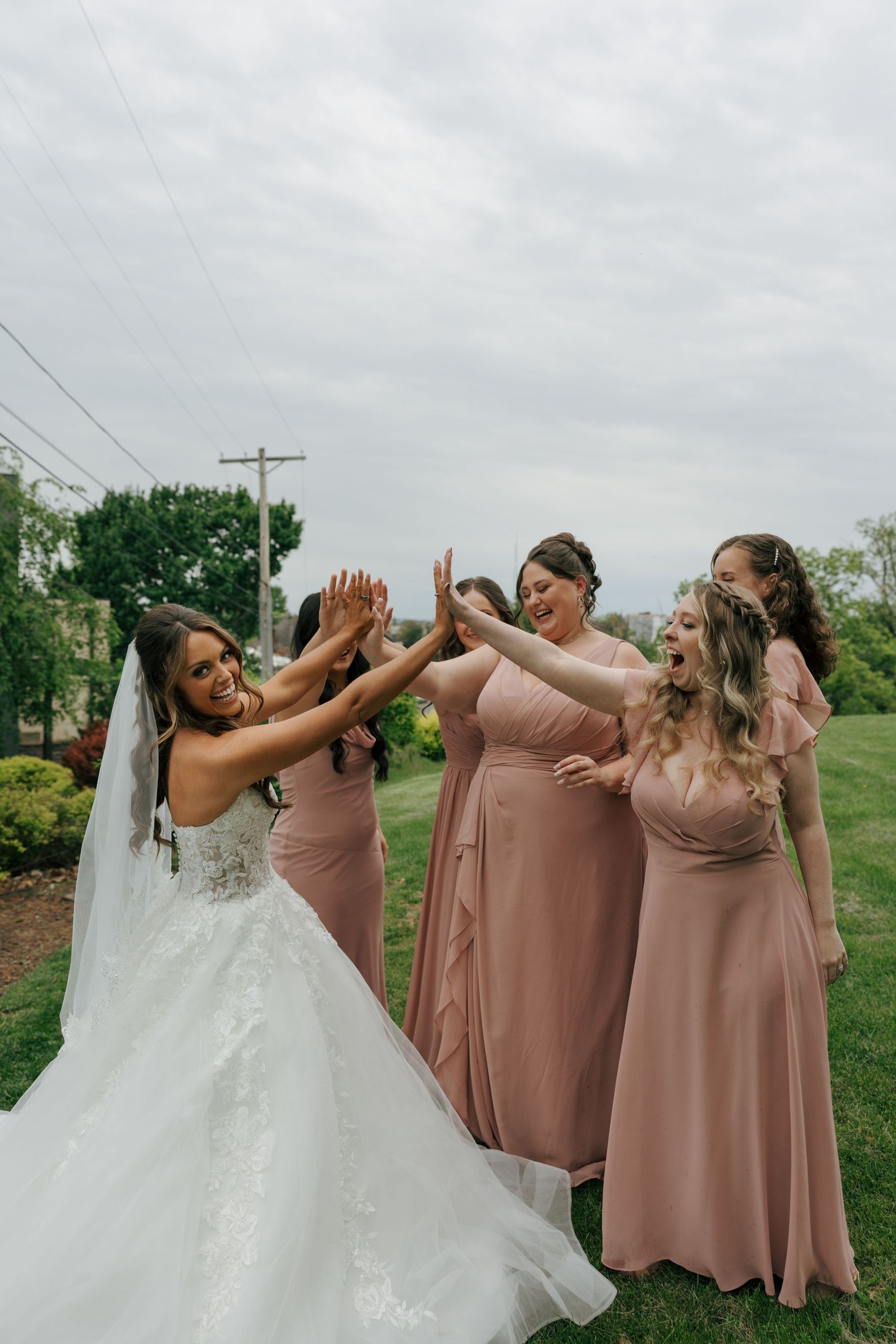 Bride in white lace gown high-fives bridesmaids in dusty rose dresses on green lawn.