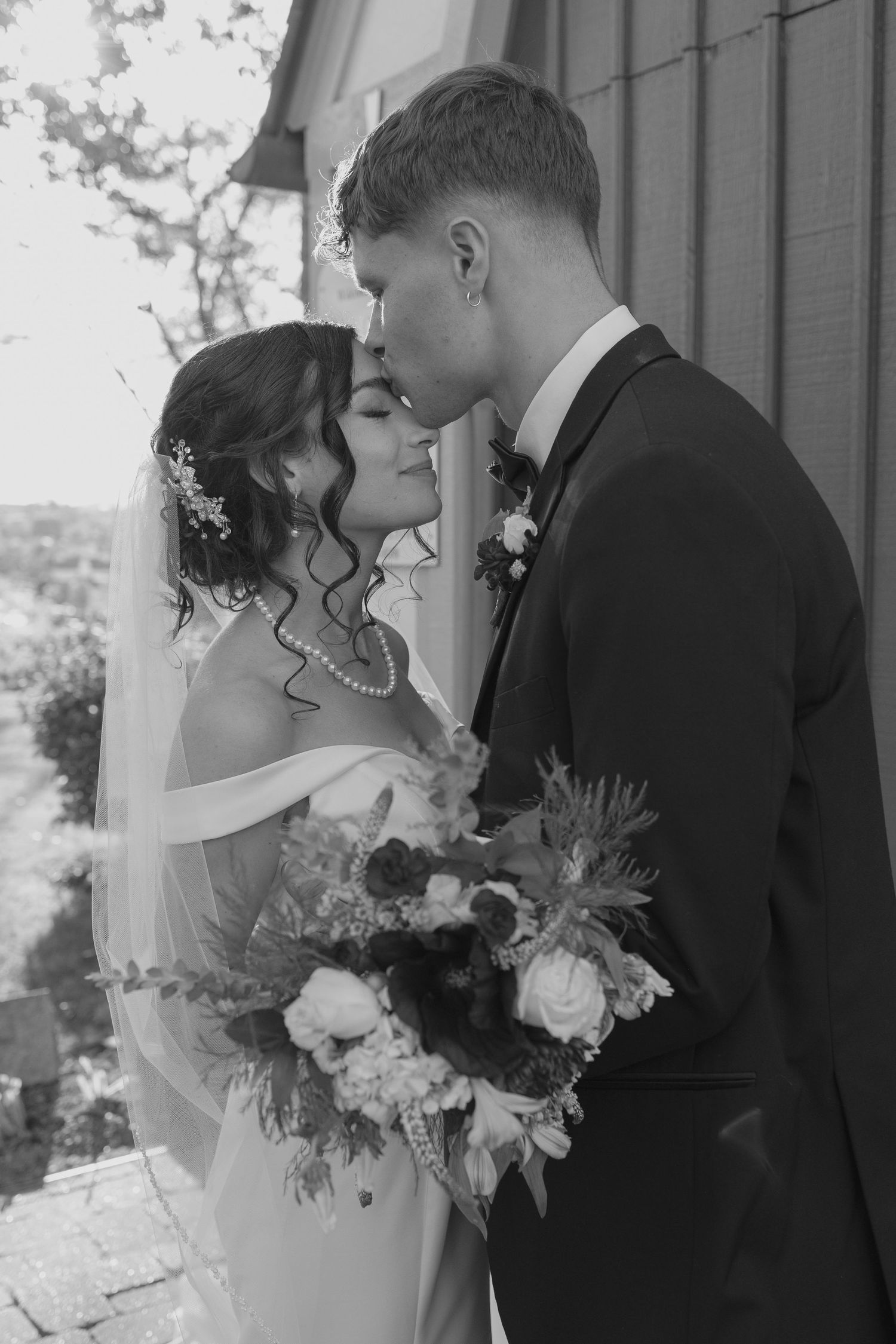Black and white portrait of groom kissing bride on forehead, bride holding large bouquet outside historic church.