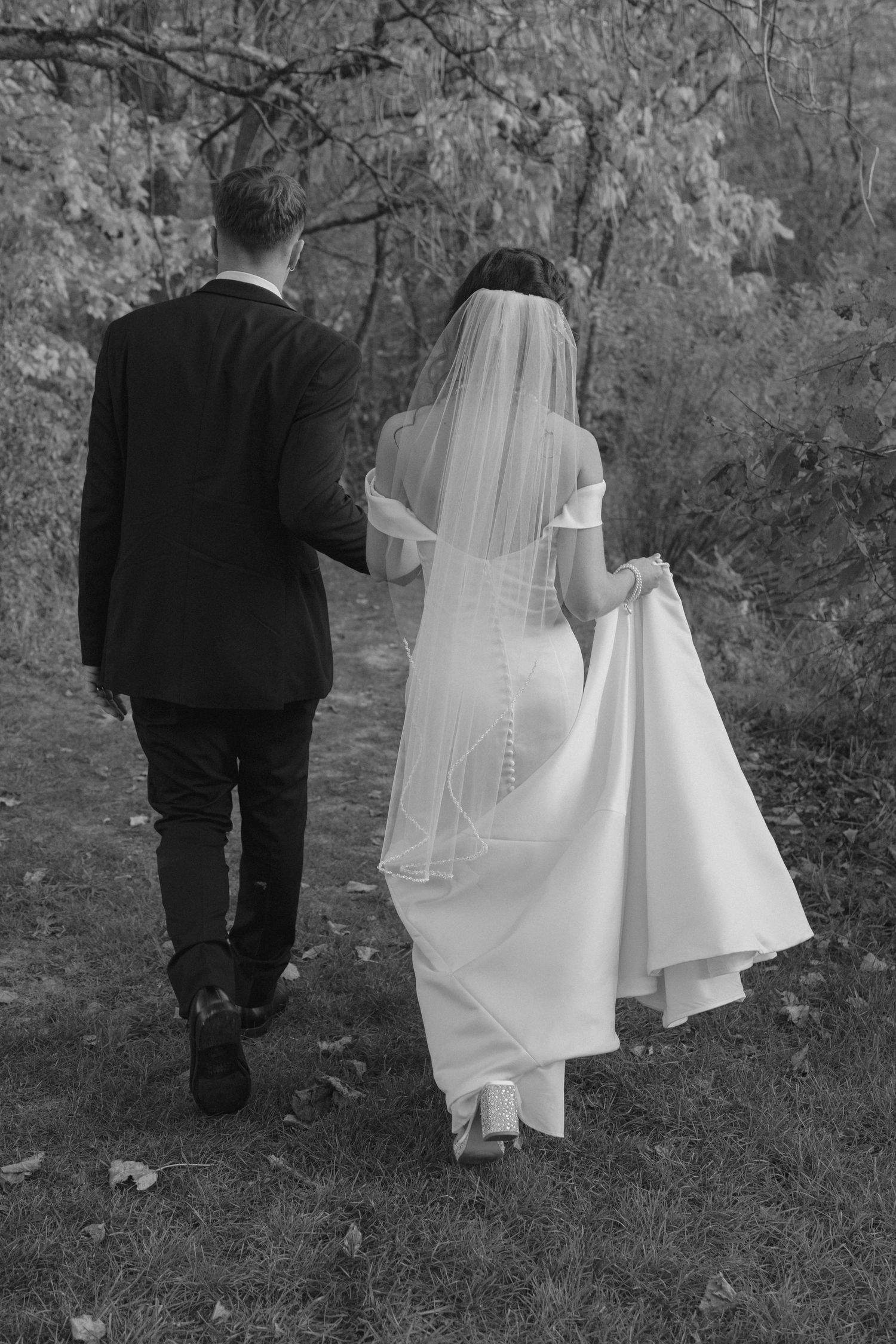 Black and white photo of bride and groom walking hand in hand along a wooded autumn path.
