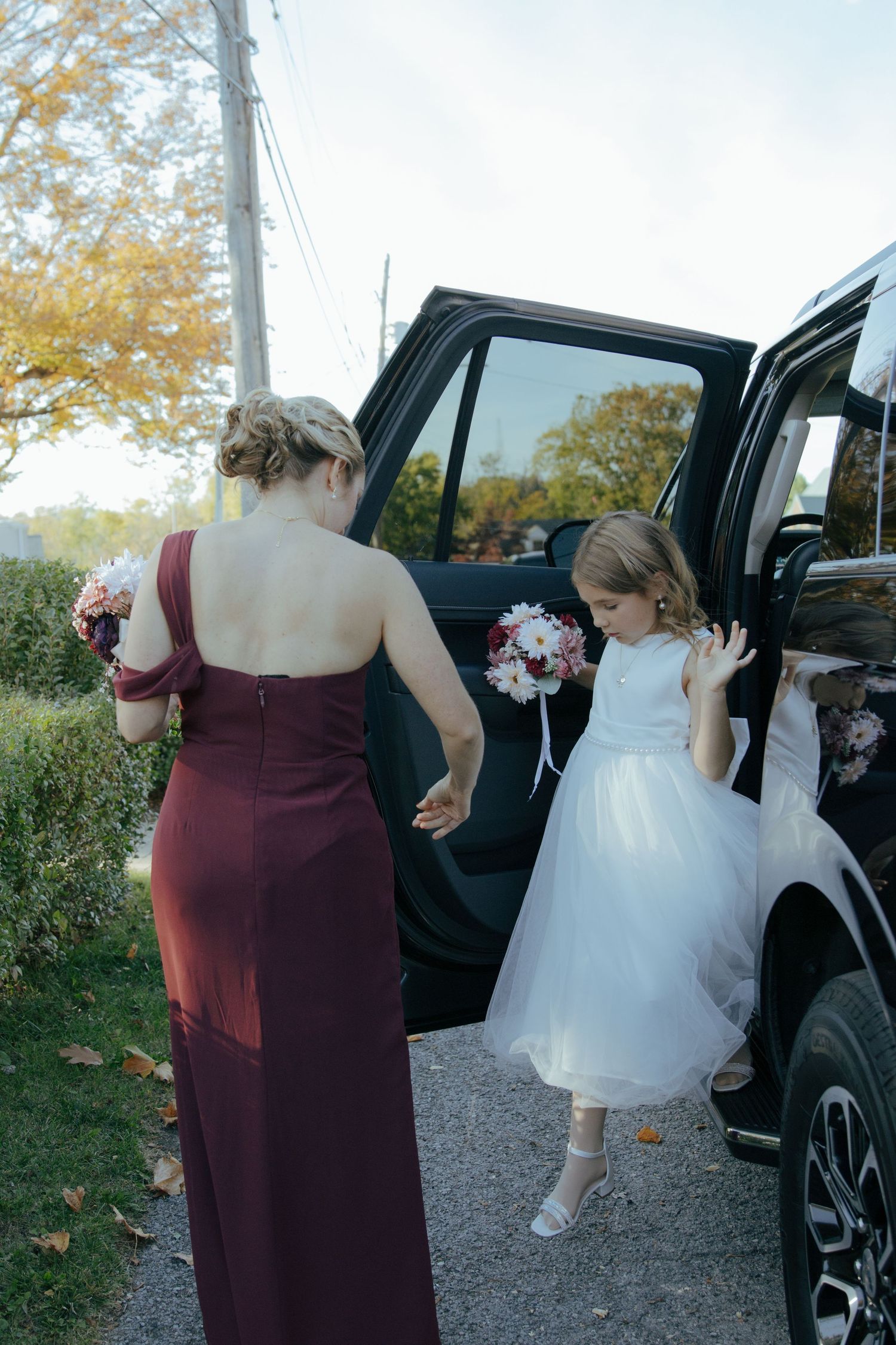 Bridesmaid in burgundy gown helps flower girl in white dress exit black SUV at outdoor fall wedding.
