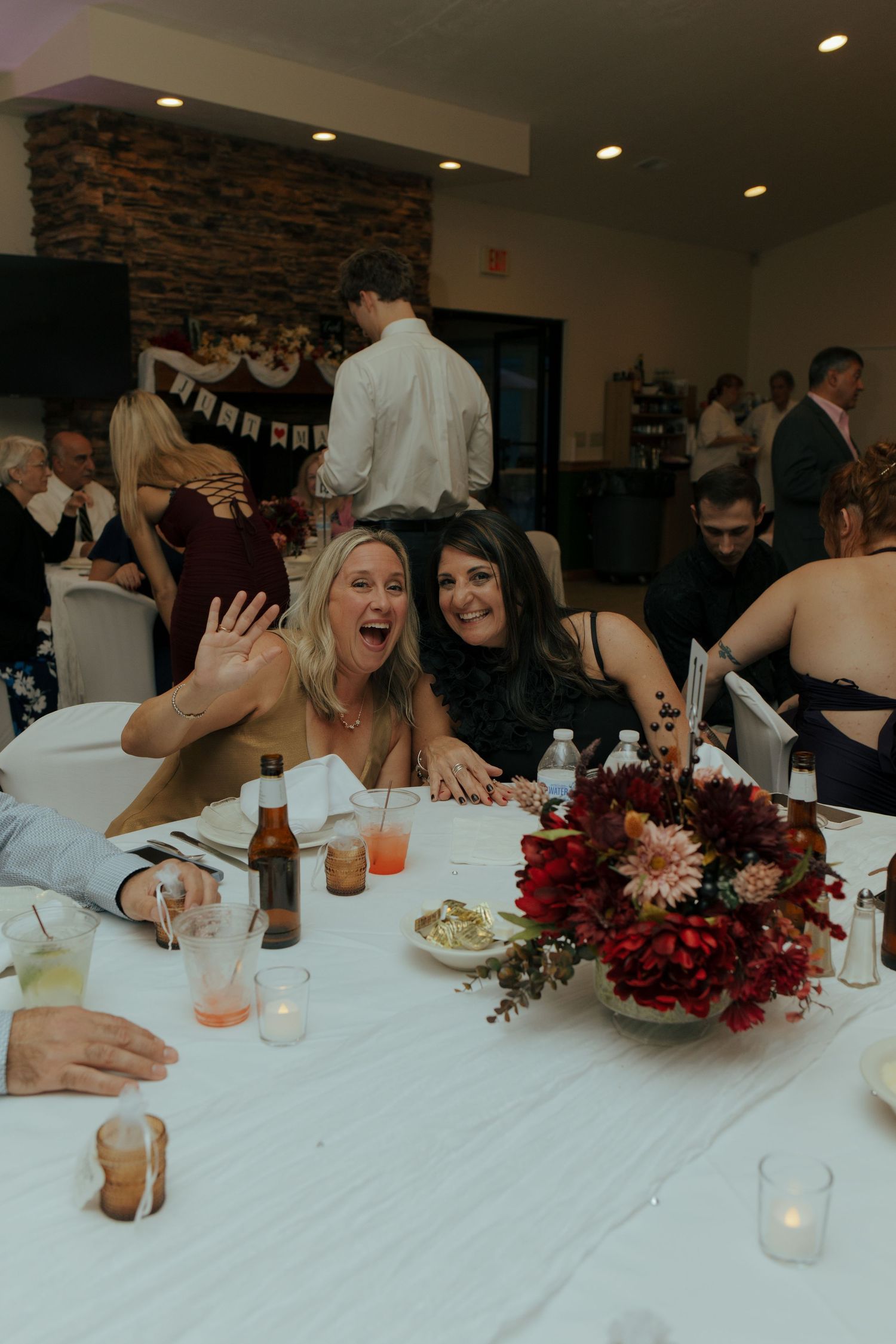 Two women smile and wave at a wedding reception table with red floral centerpiece and candles.