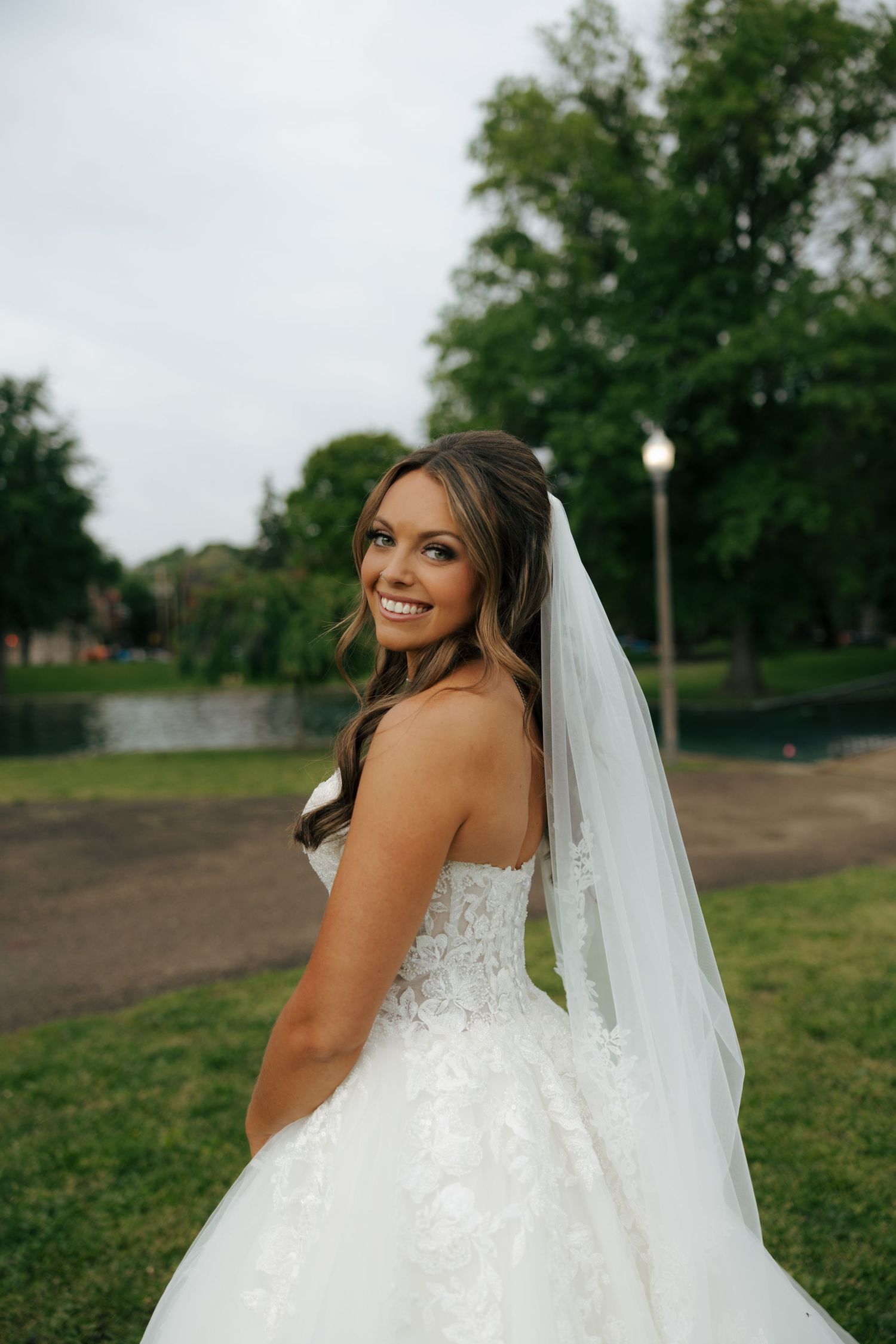 Smiling bride in a strapless lace wedding gown and veil poses outdoors in a green park setting.