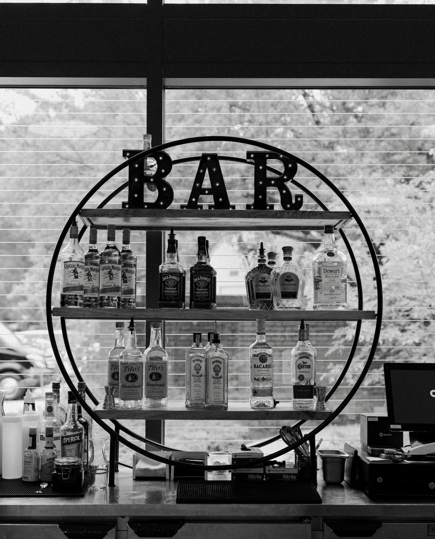 Black and white photo of a circular bar shelf display with liquor bottles and a marquee BAR sign, set against a window.