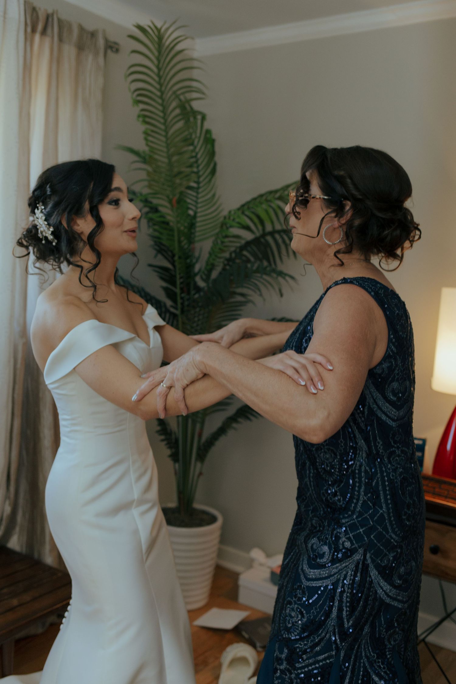 Bride in off-shoulder white gown holds hands with mother in navy beaded dress while getting ready.
