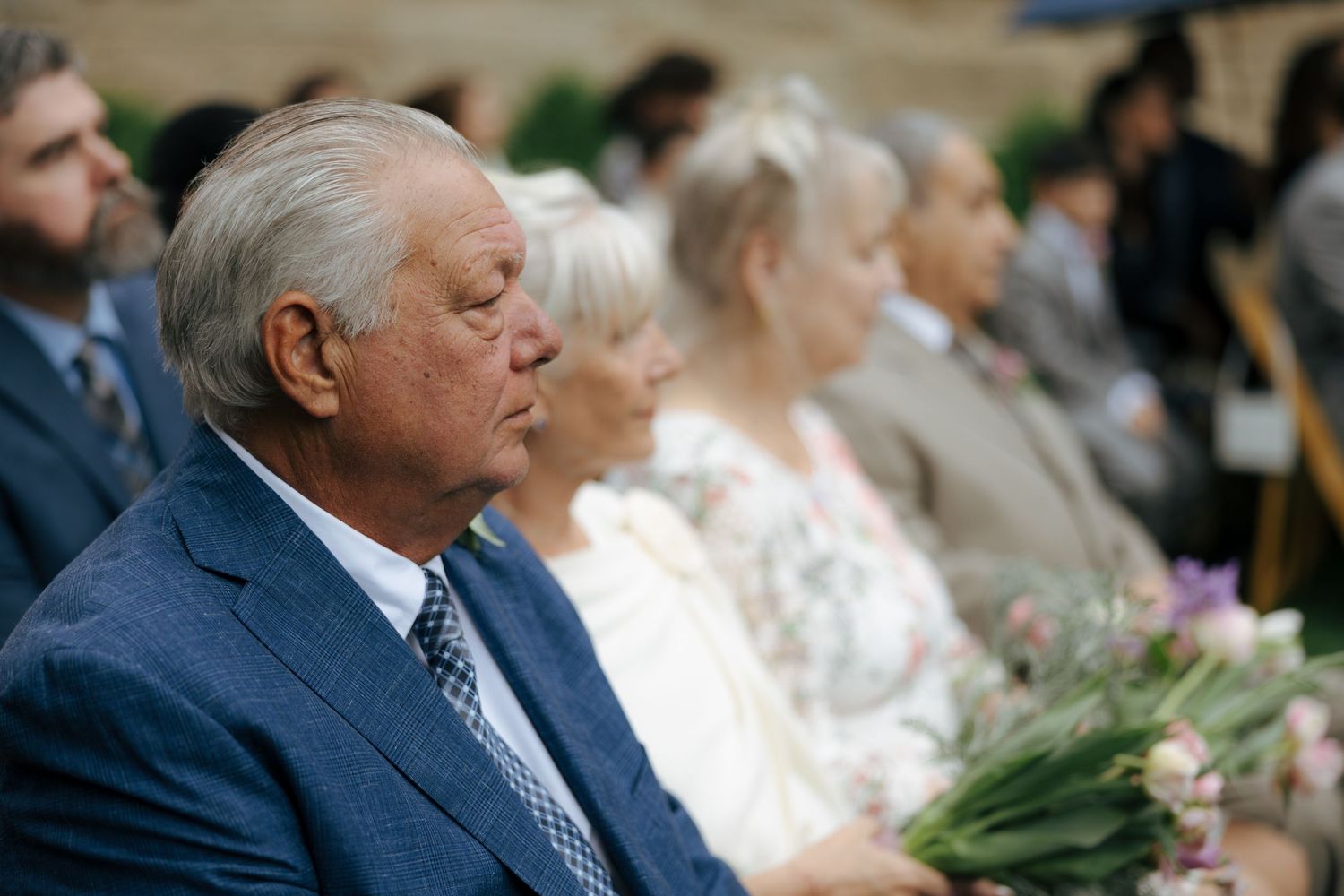 Elderly man in blue suit seated among wedding guests during outdoor ceremony.