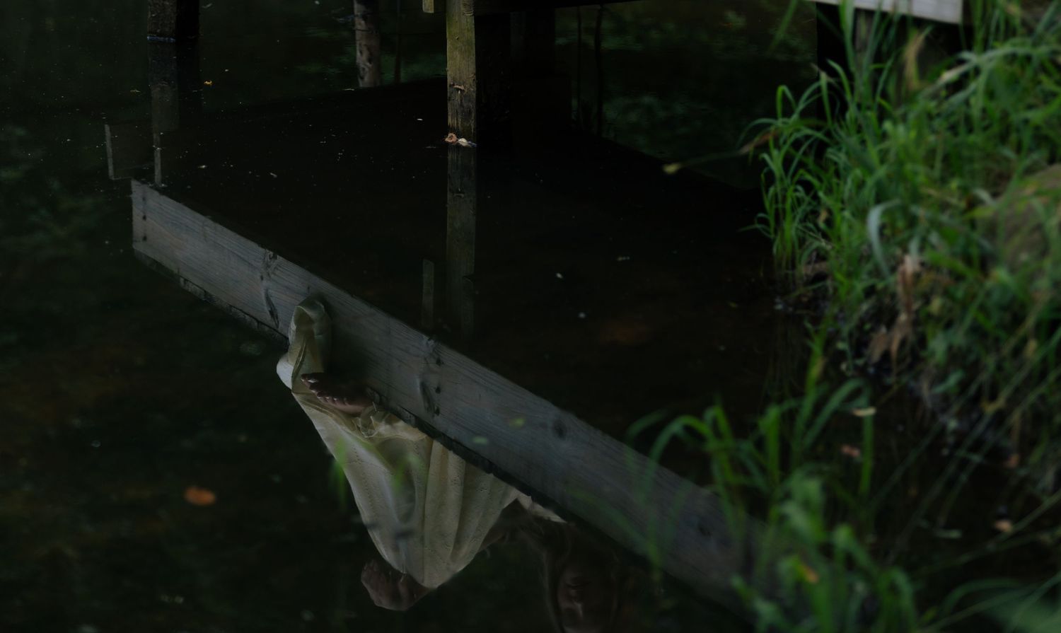 Reflection of wooden dock and fabric in dark calm water surrounded by greenery.