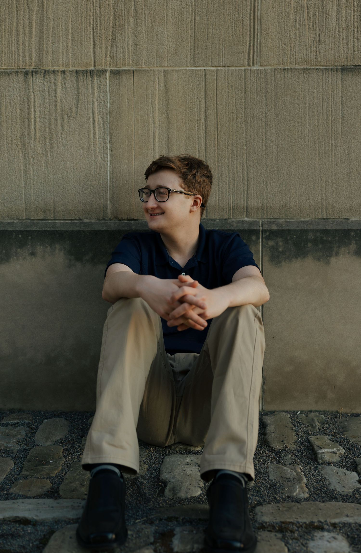 Teen boy in glasses and khakis sits against stone wall on cobblestone ground.