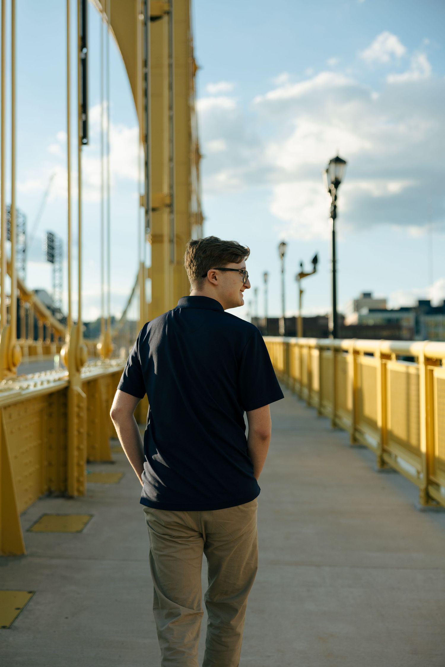 Young man in navy t-shirt and khakis walks on yellow bridge, viewed from behind, city skyline visible.