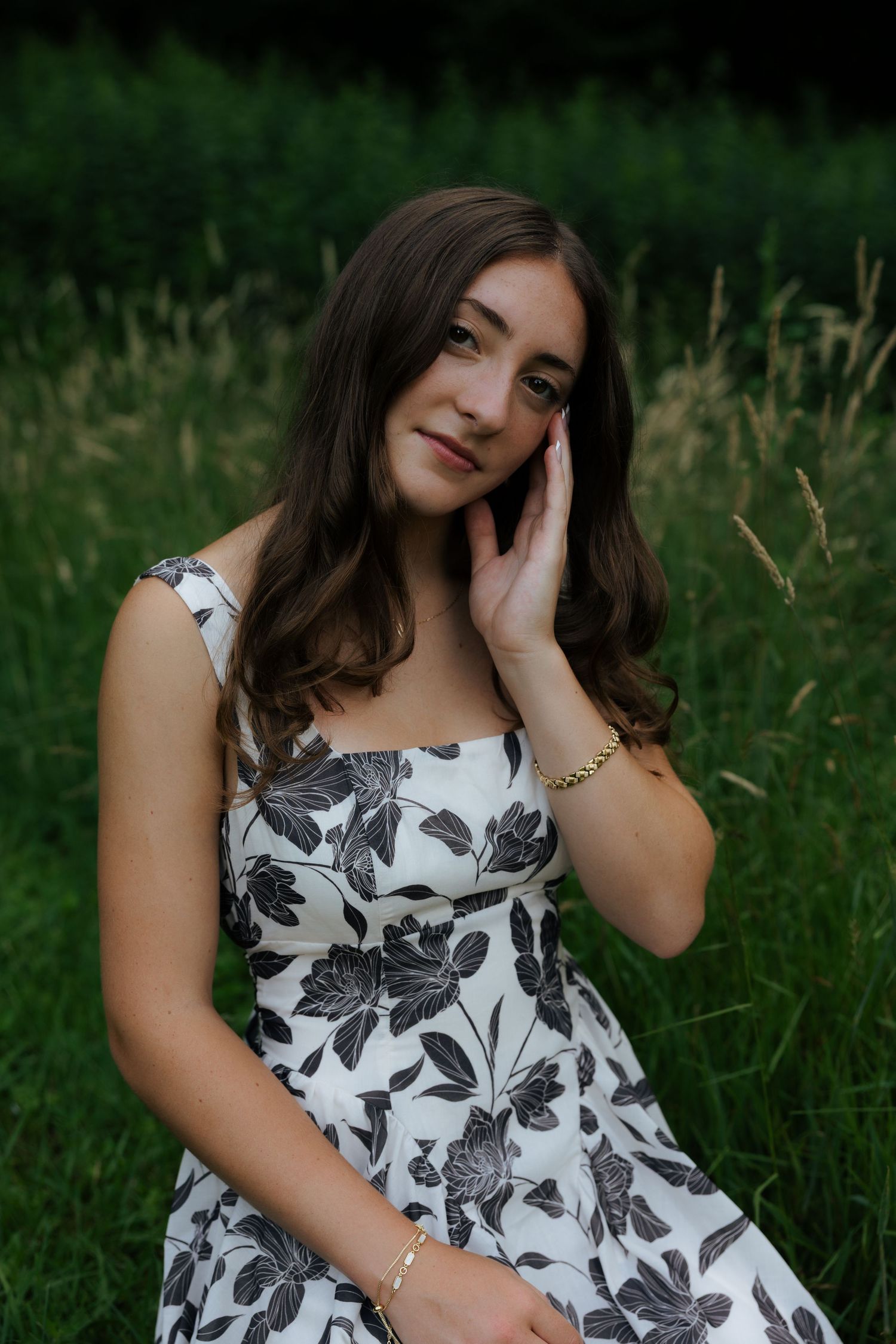 A young woman in a black and white floral dress sits in tall grass, gently touching her face.