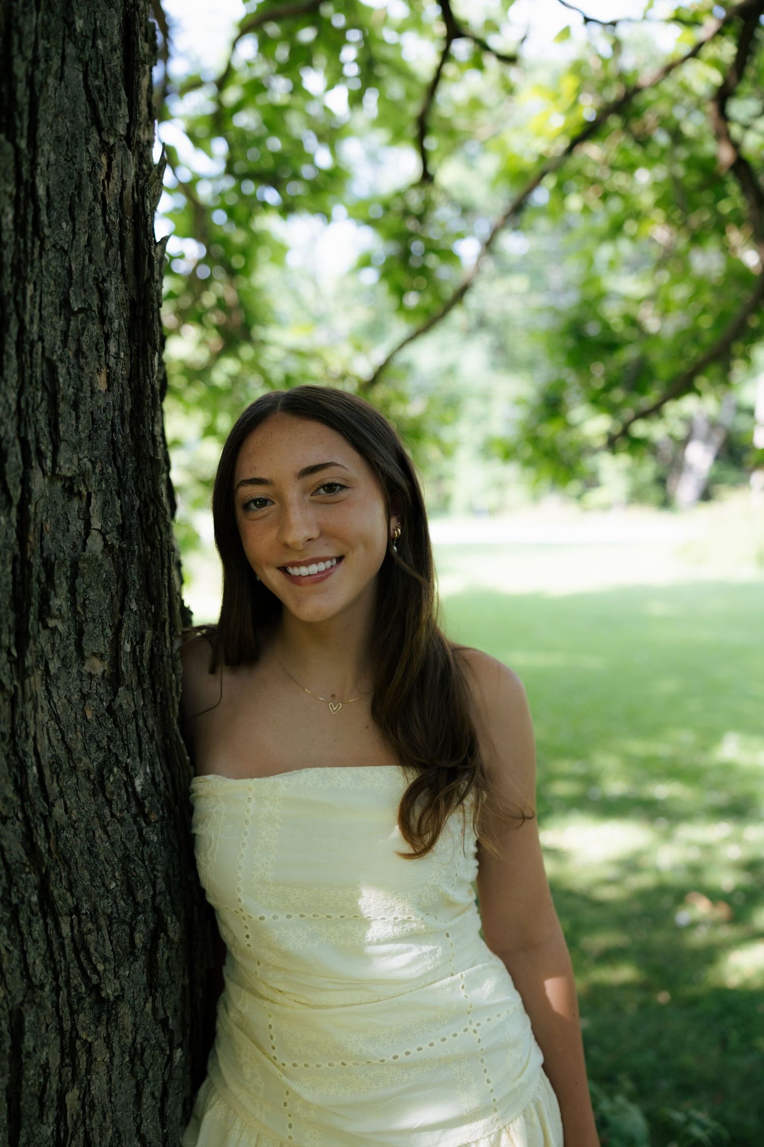 Smiling young woman in white strapless dress leaning against tree trunk in sunny green park.