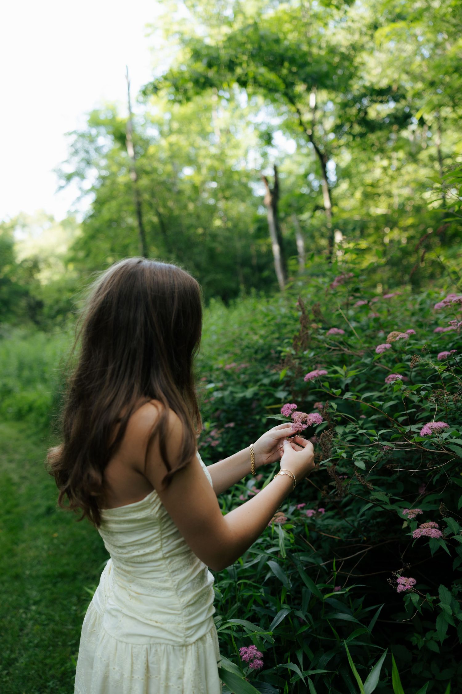 Young woman in cream dress reaching to touch pink flowers on bush along forest path.