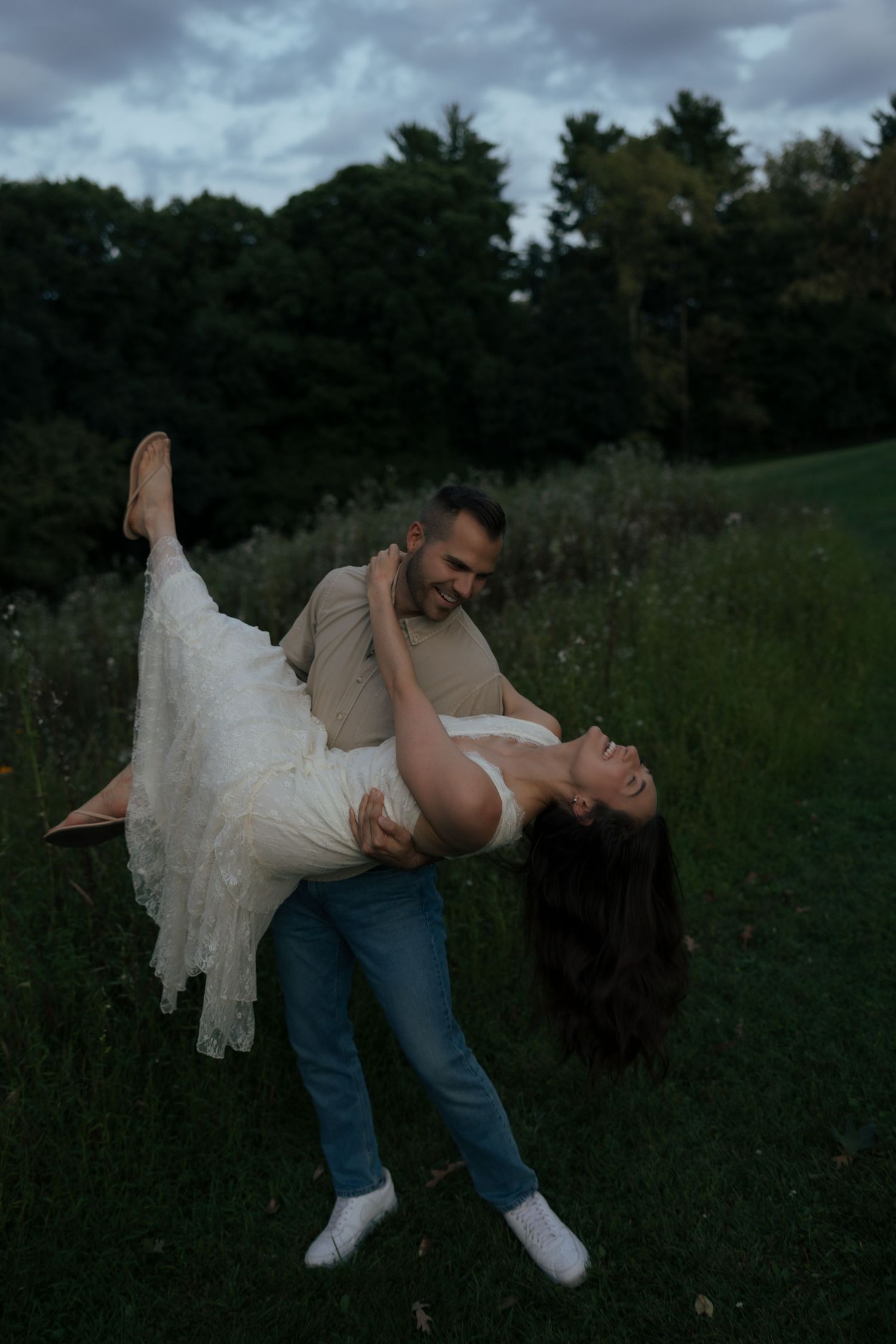 Man in jeans lifting and spinning laughing woman in white dress in a green field at dusk.