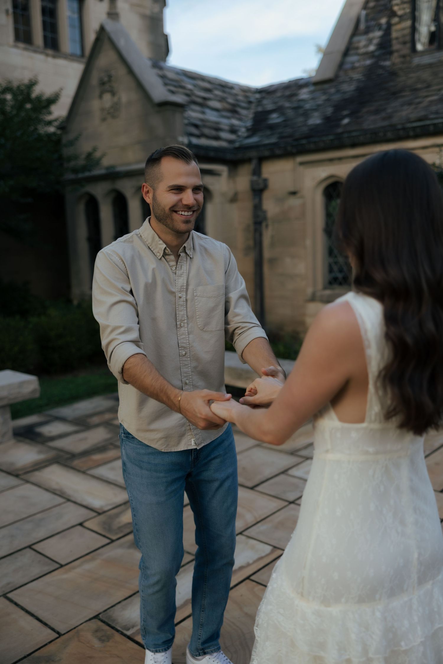 Smiling man holds woman's hands during outdoor marriage proposal at a historic stone mansion.
