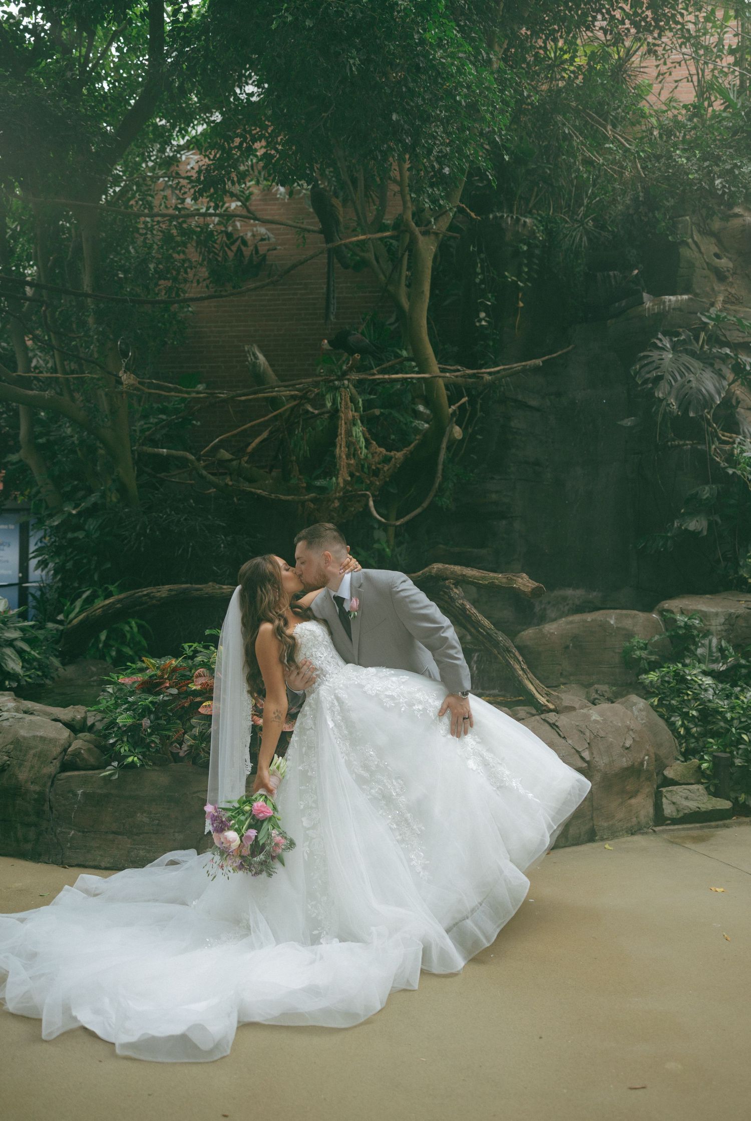 A bride and groom share a romantic kiss in a lush garden setting, with the bride wearing a flowing white ballgown and veil.