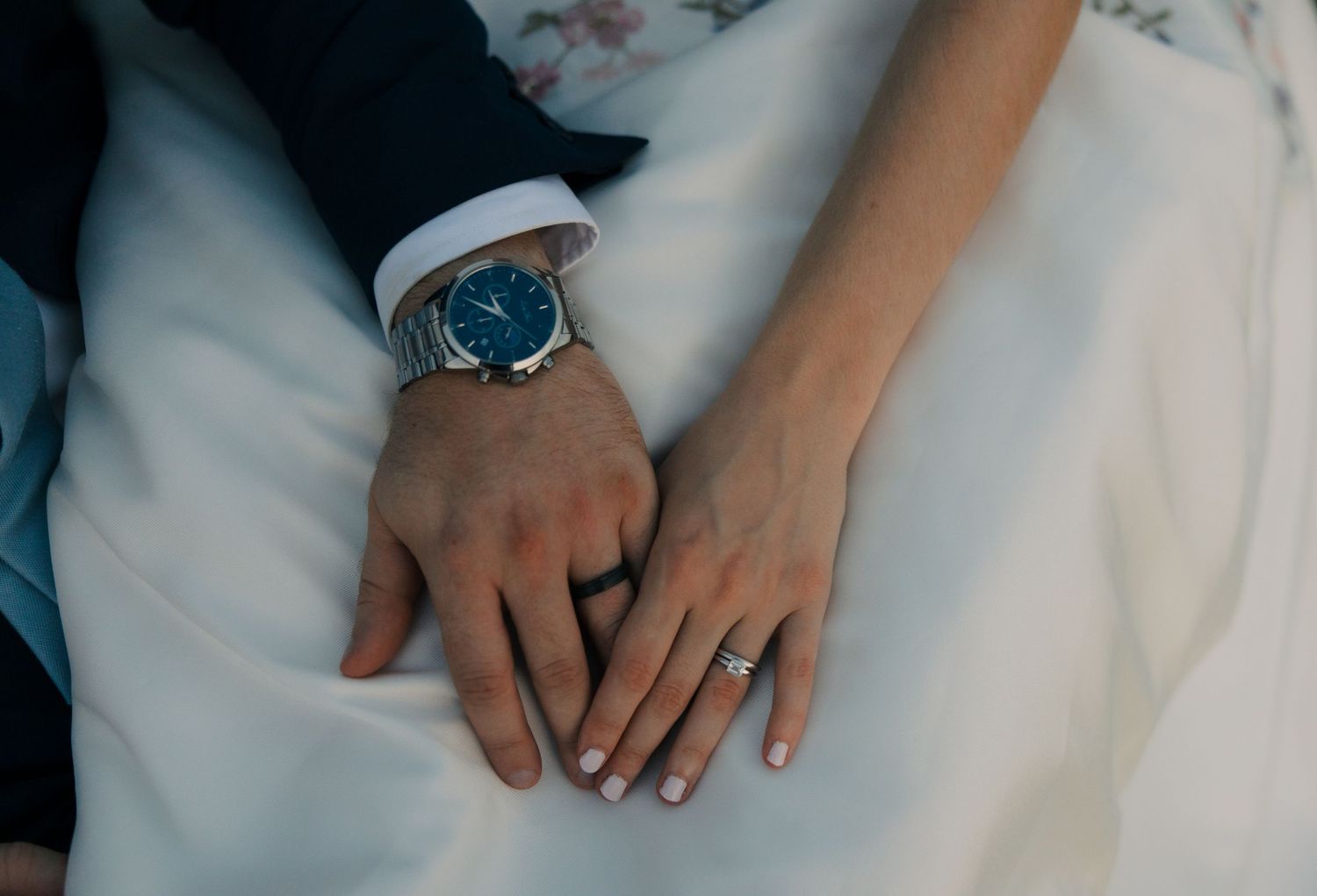 Close-up of newlyweds hands showing a diamond engagement ring and a blue-faced dress watch on white fabric.
