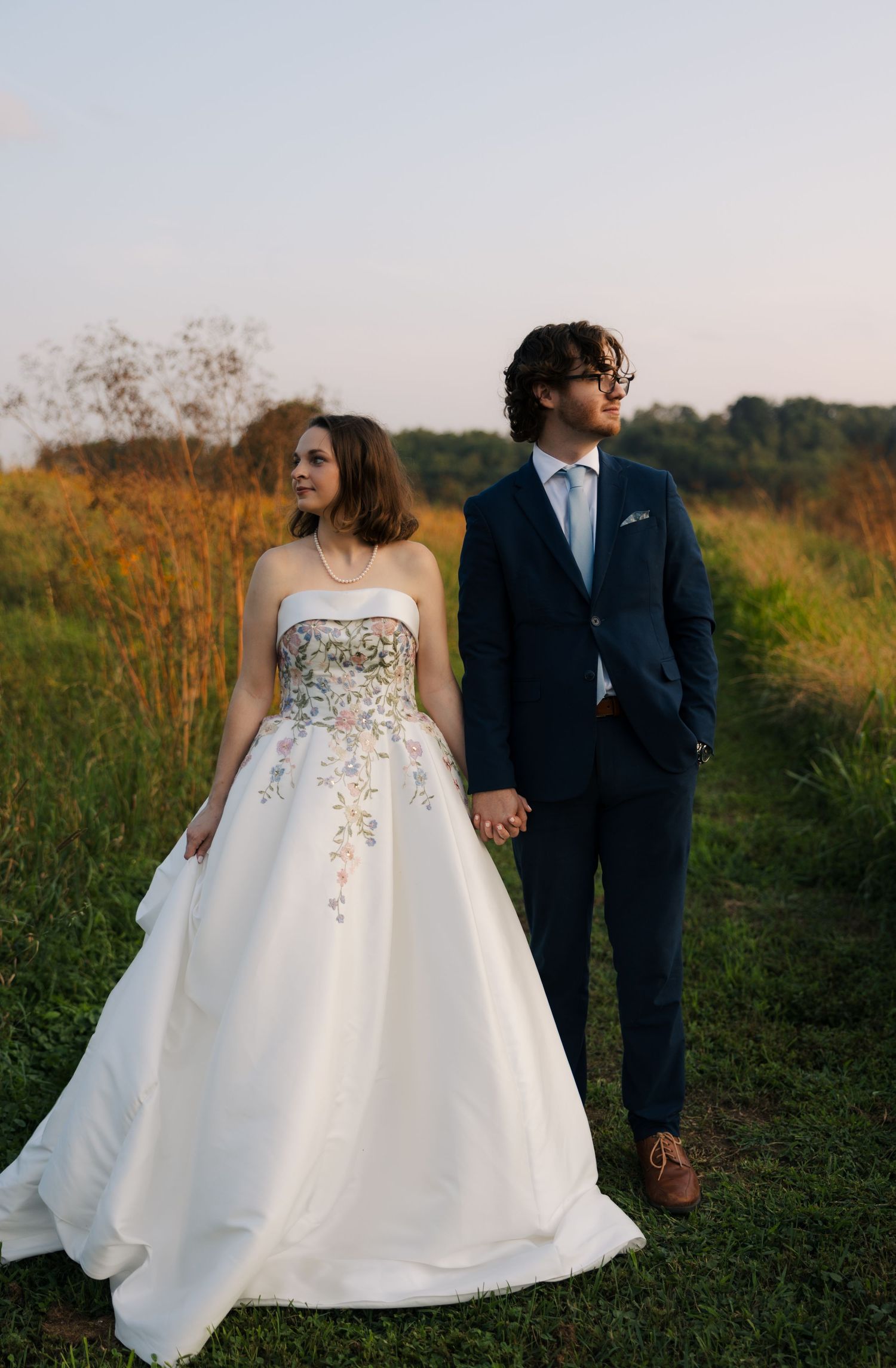 A bride in a floral strapless ball gown holds hands with a groom in a navy suit in a sunlit field.