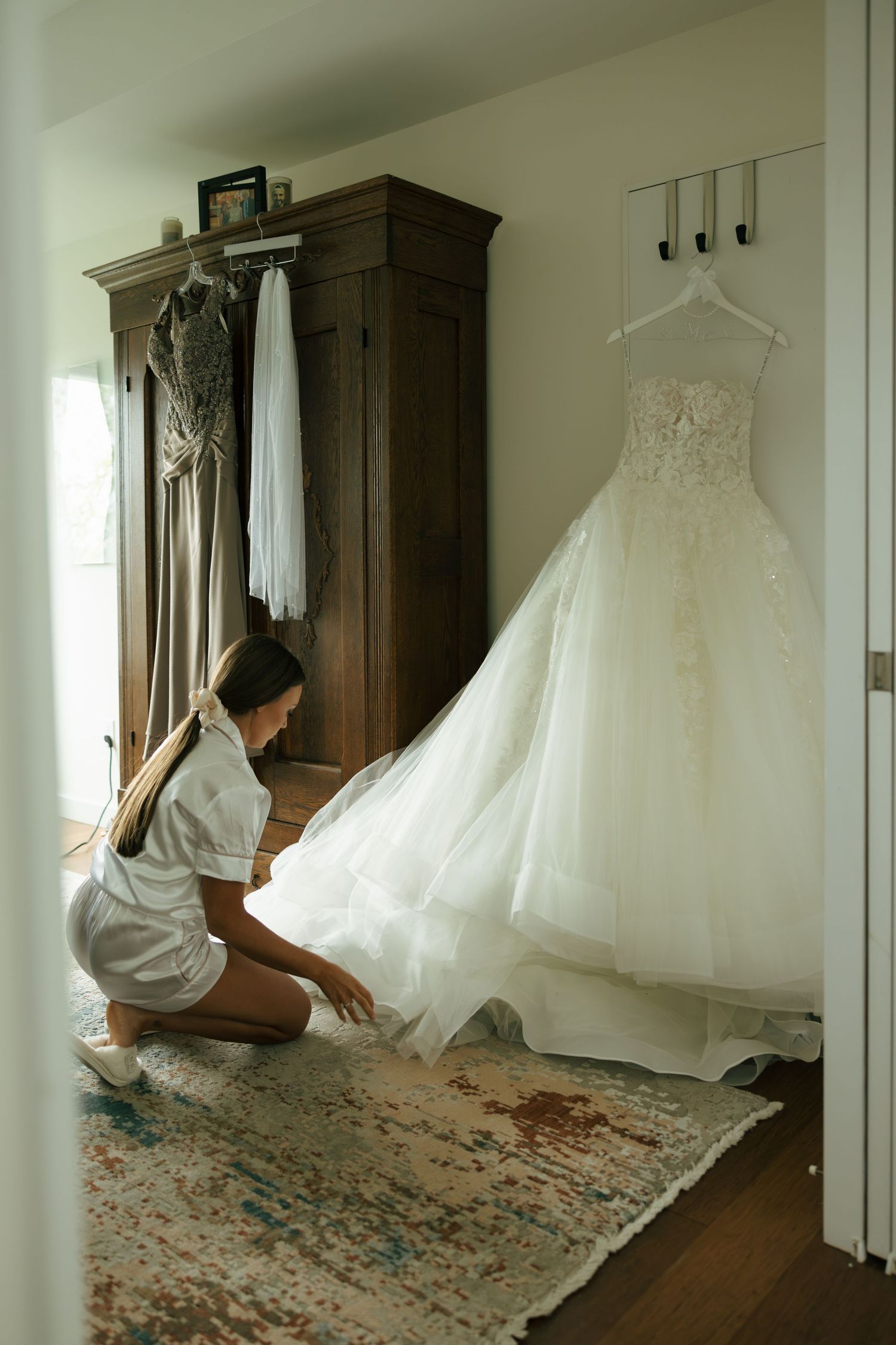 Bride kneeling beside her elegant lace wedding gown hanging in a cozy room with a wooden armoire.