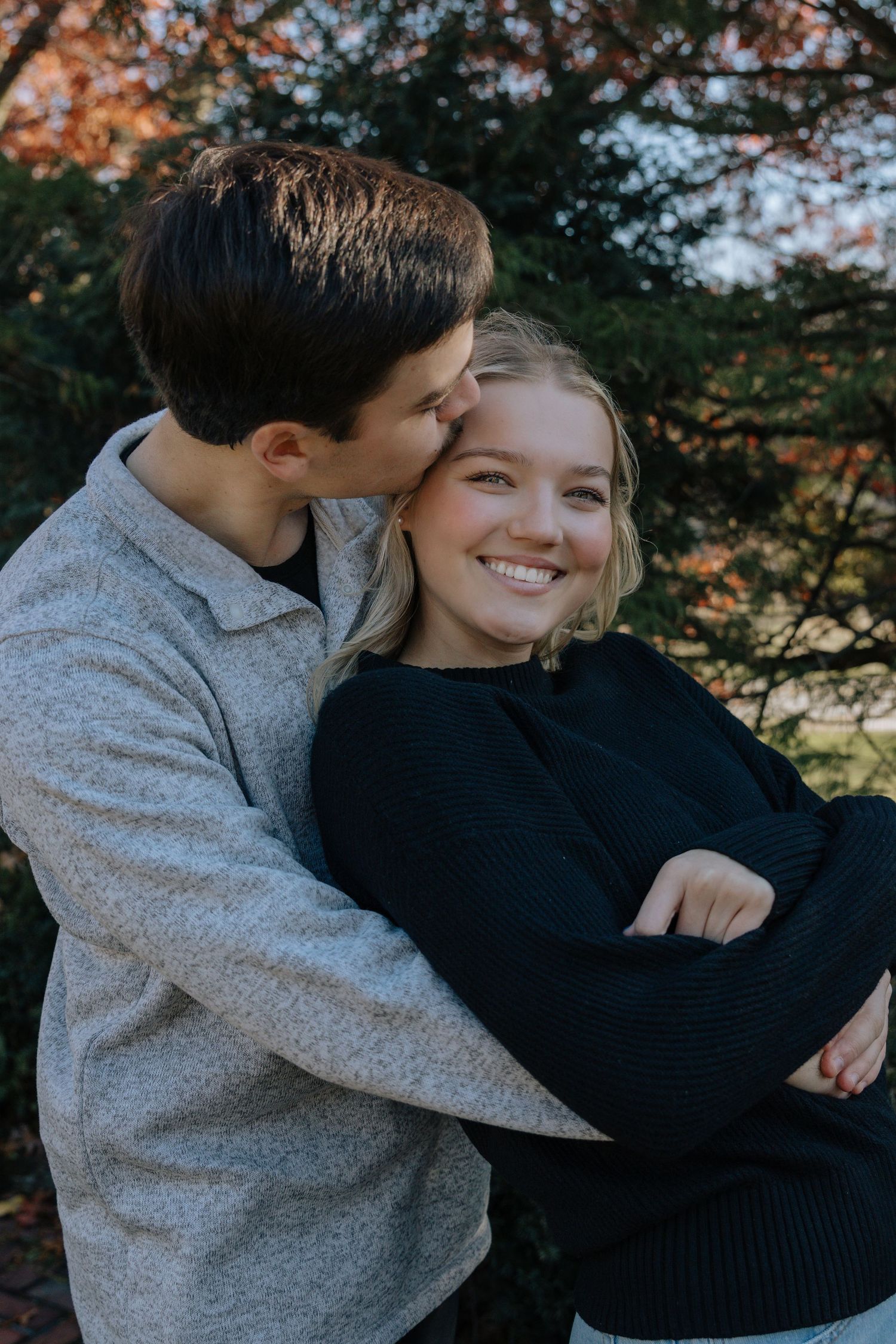 A couple shares an intimate moment outdoors during autumn with fall foliage in the background.