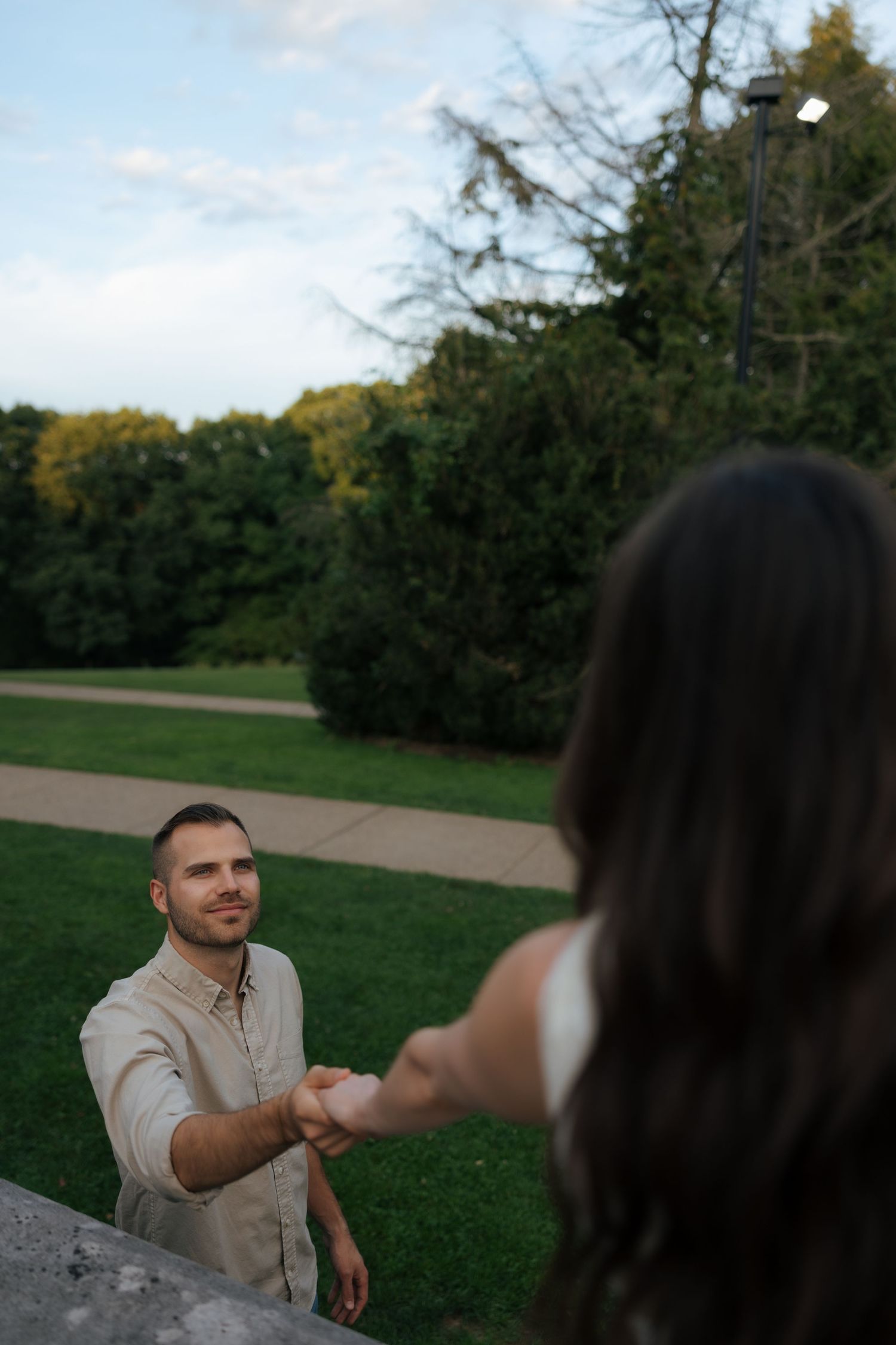 A smiling man in a beige shirt holds hands with a woman in a lush green park setting.
