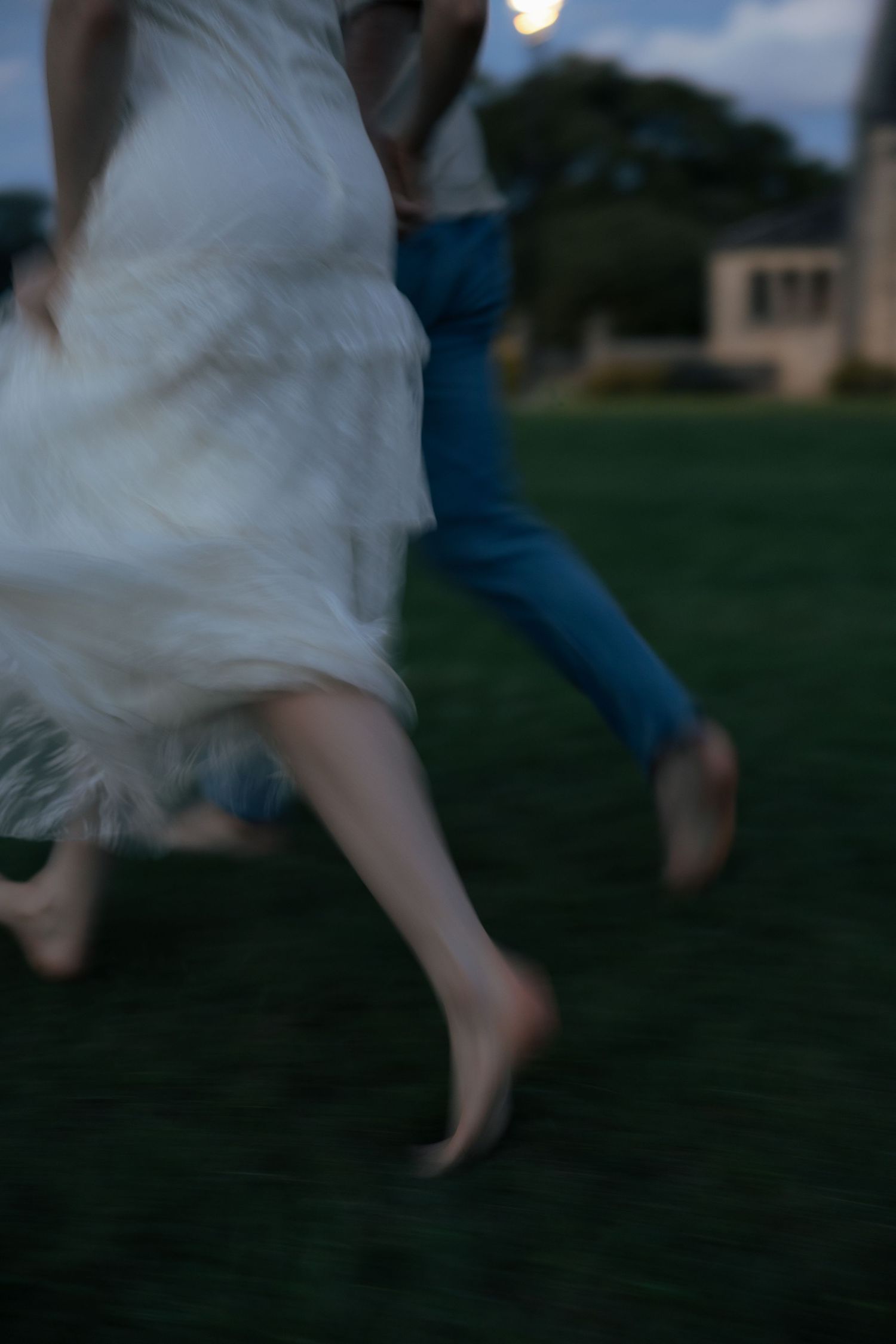 Motion blur shot of couple running barefoot on grass at dusk in white dress and jeans.