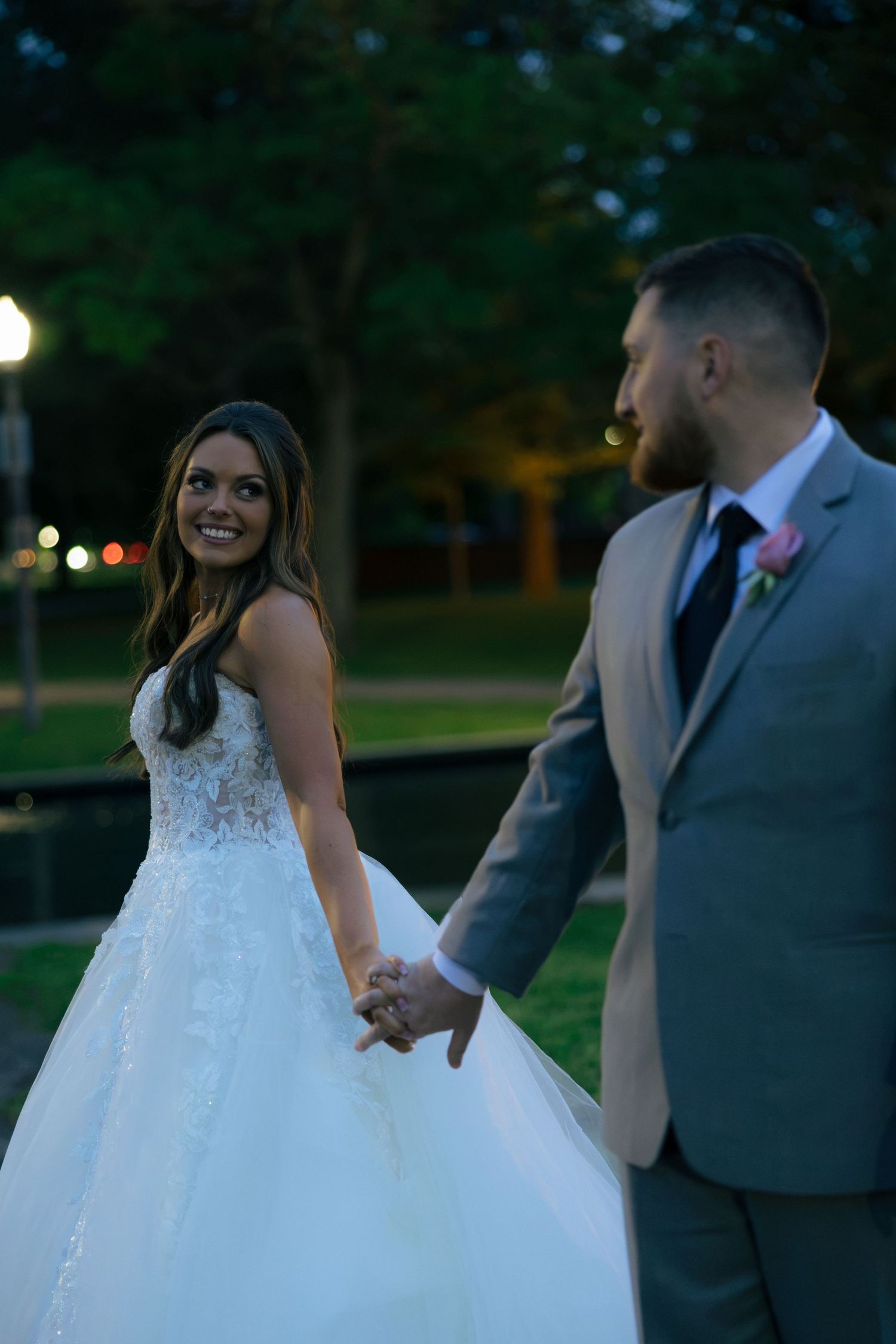 Bride in white lace ball gown holds hands with groom in gray suit outdoors at dusk, smiling joyfully.