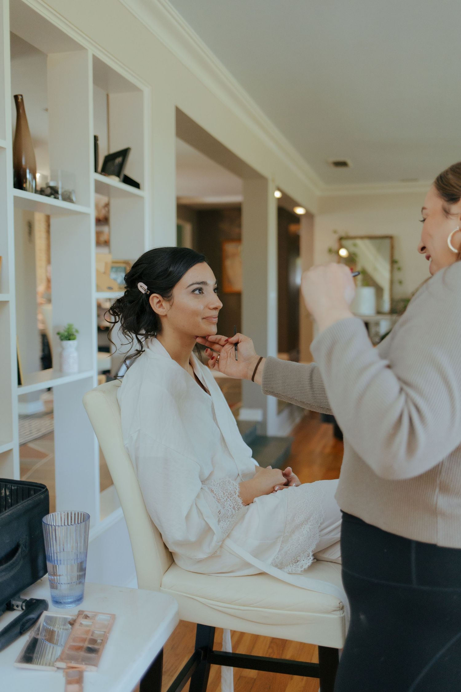 Bride in white robe getting makeup applied by professional artist in a bright, elegant room.