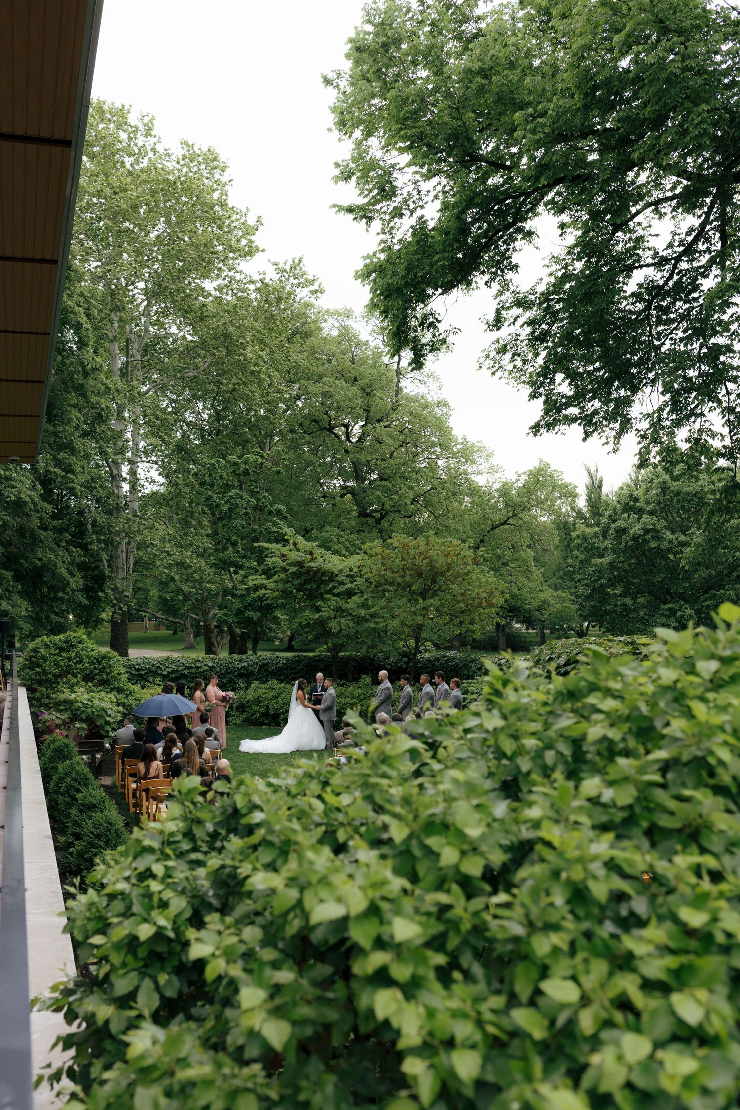 Aerial view of outdoor garden wedding ceremony surrounded by lush green trees and hedges.