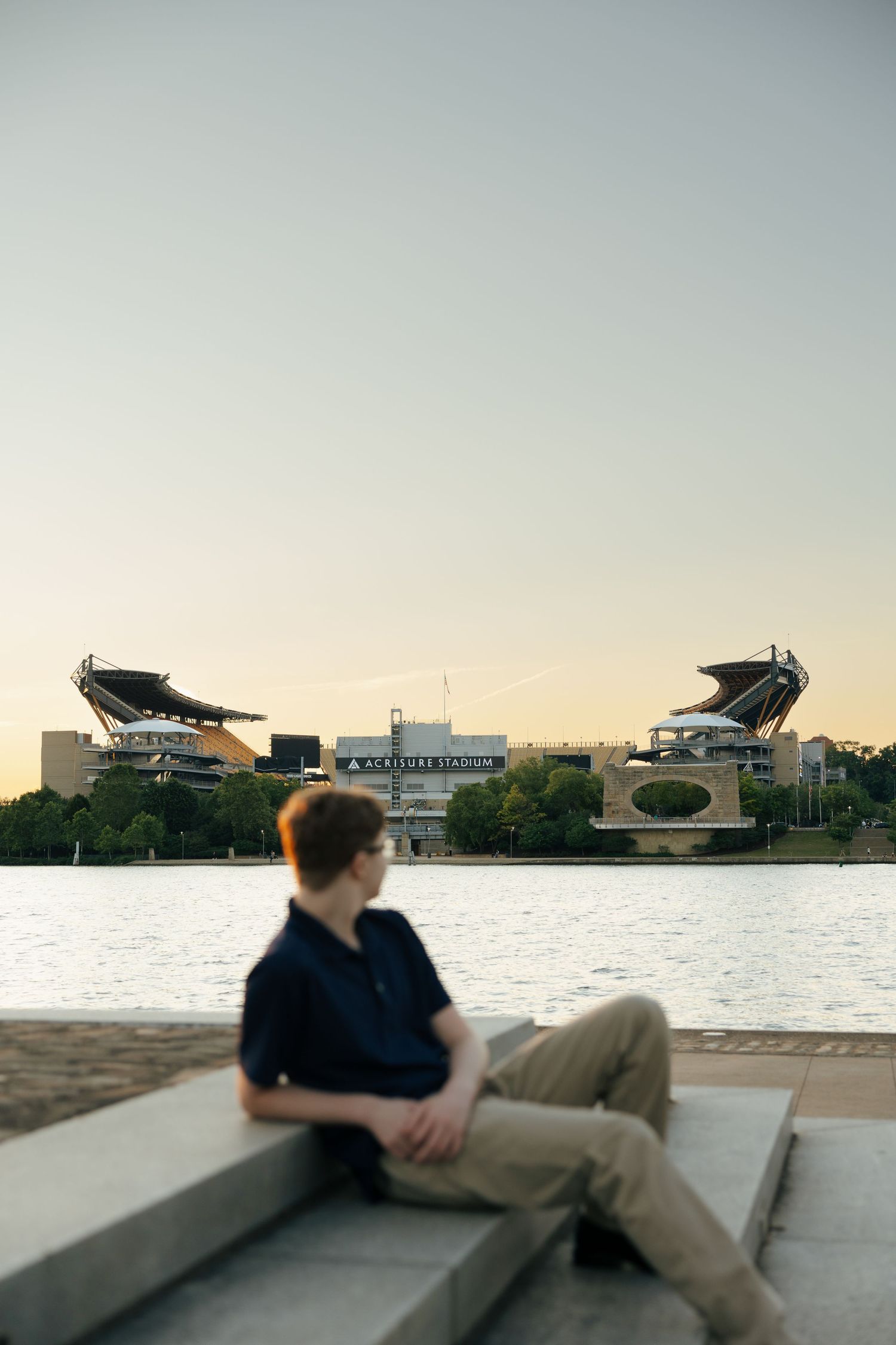 Young man sitting on steps by a river with Acrisure Stadium in the background at sunset.