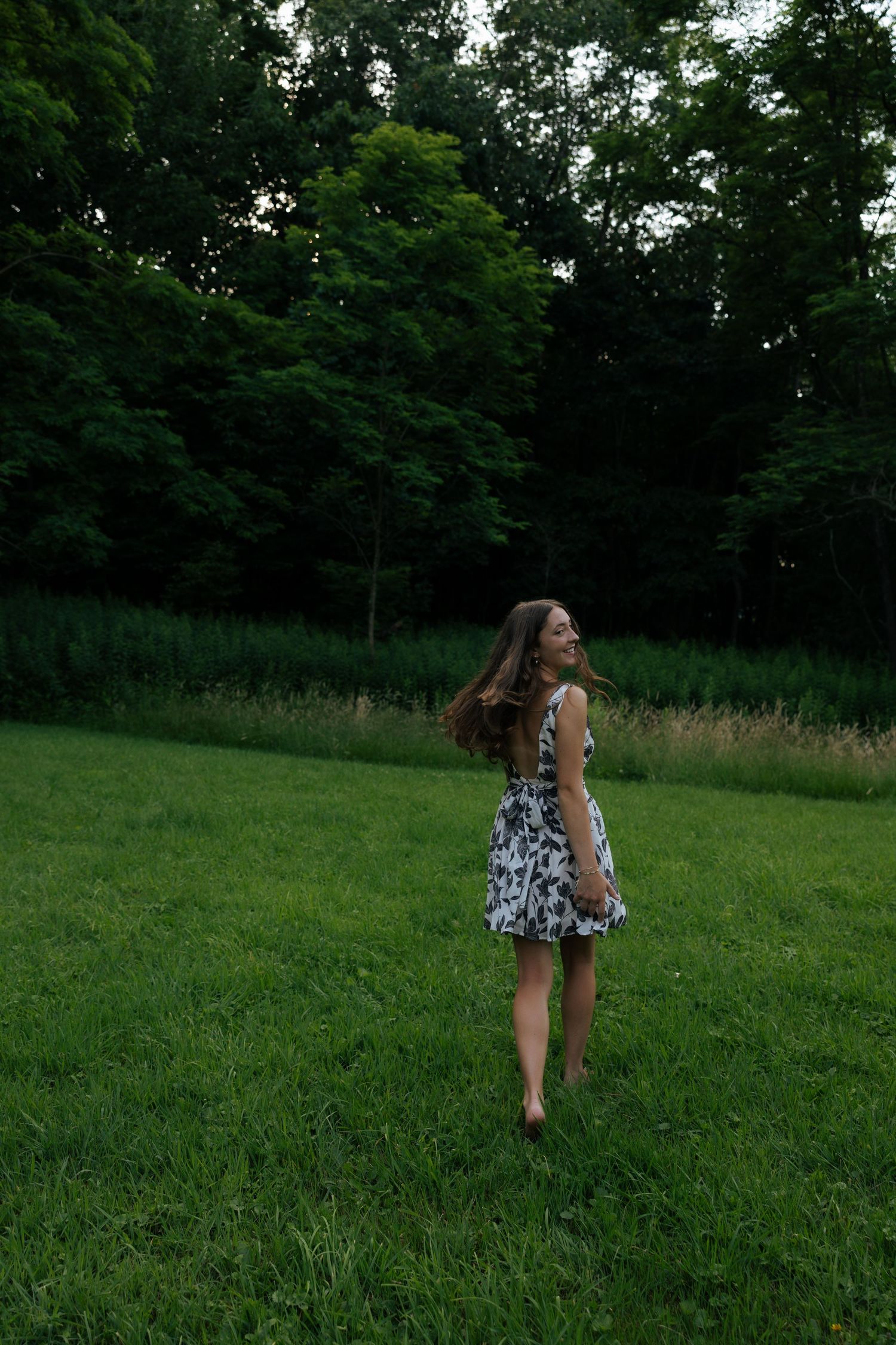 Young woman in black and white floral dress twirling barefoot on lush green lawn with trees behind.
