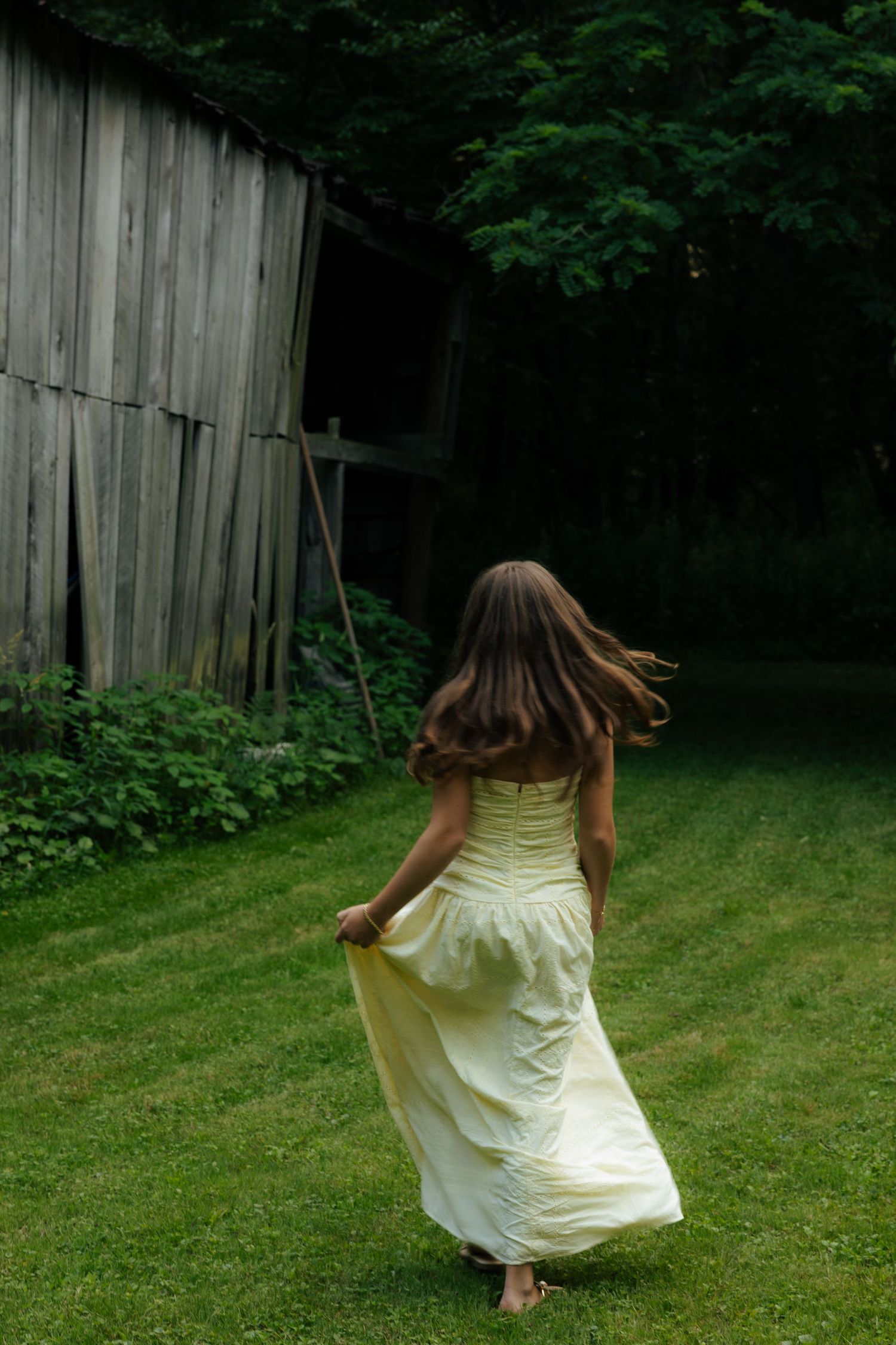 Young woman in flowing white dress running barefoot toward rustic barn in lush green landscape.
