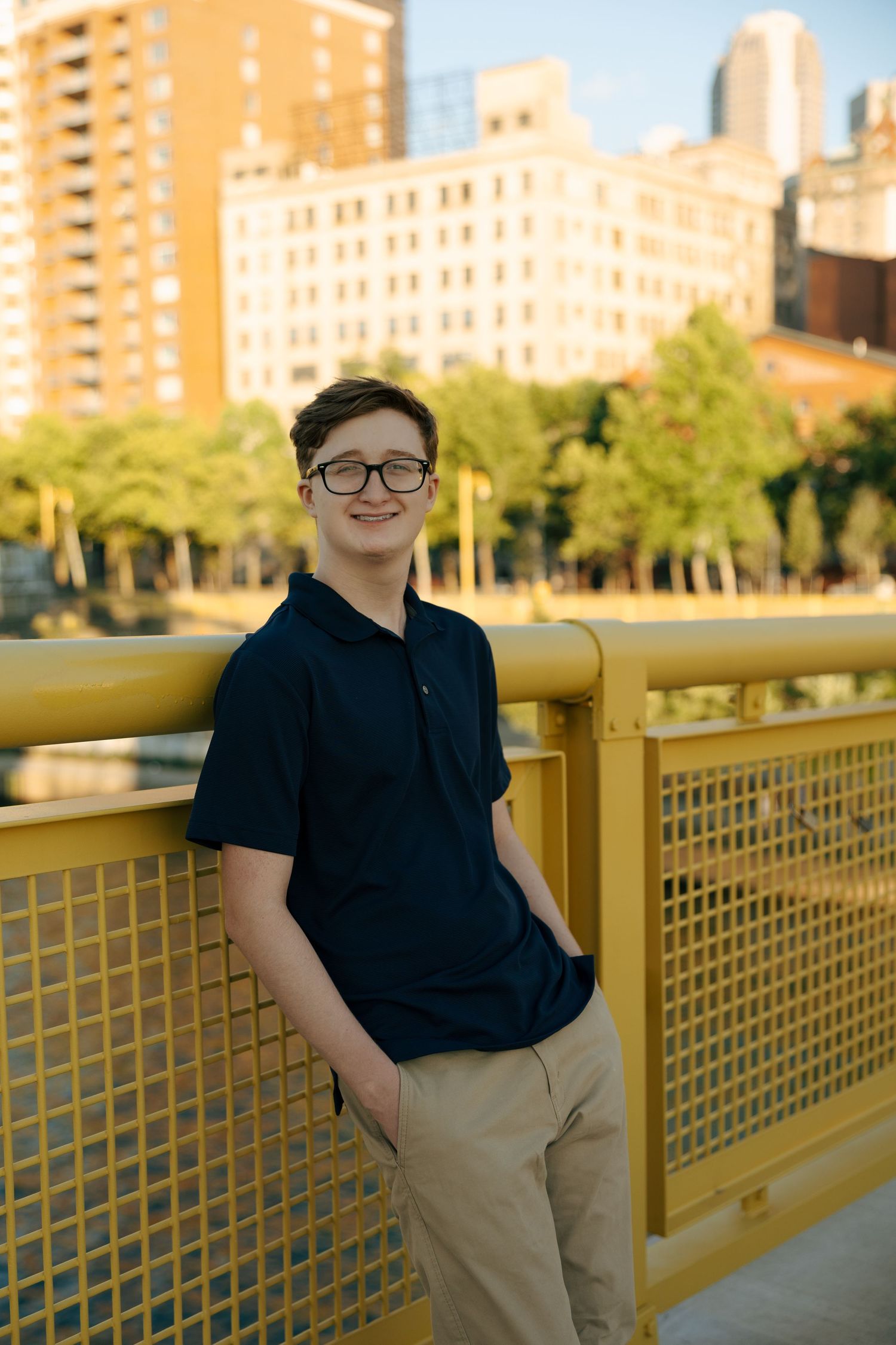 Smiling teenage boy in navy polo shirt posing on yellow Pittsburgh bridge with city skyline behind him.